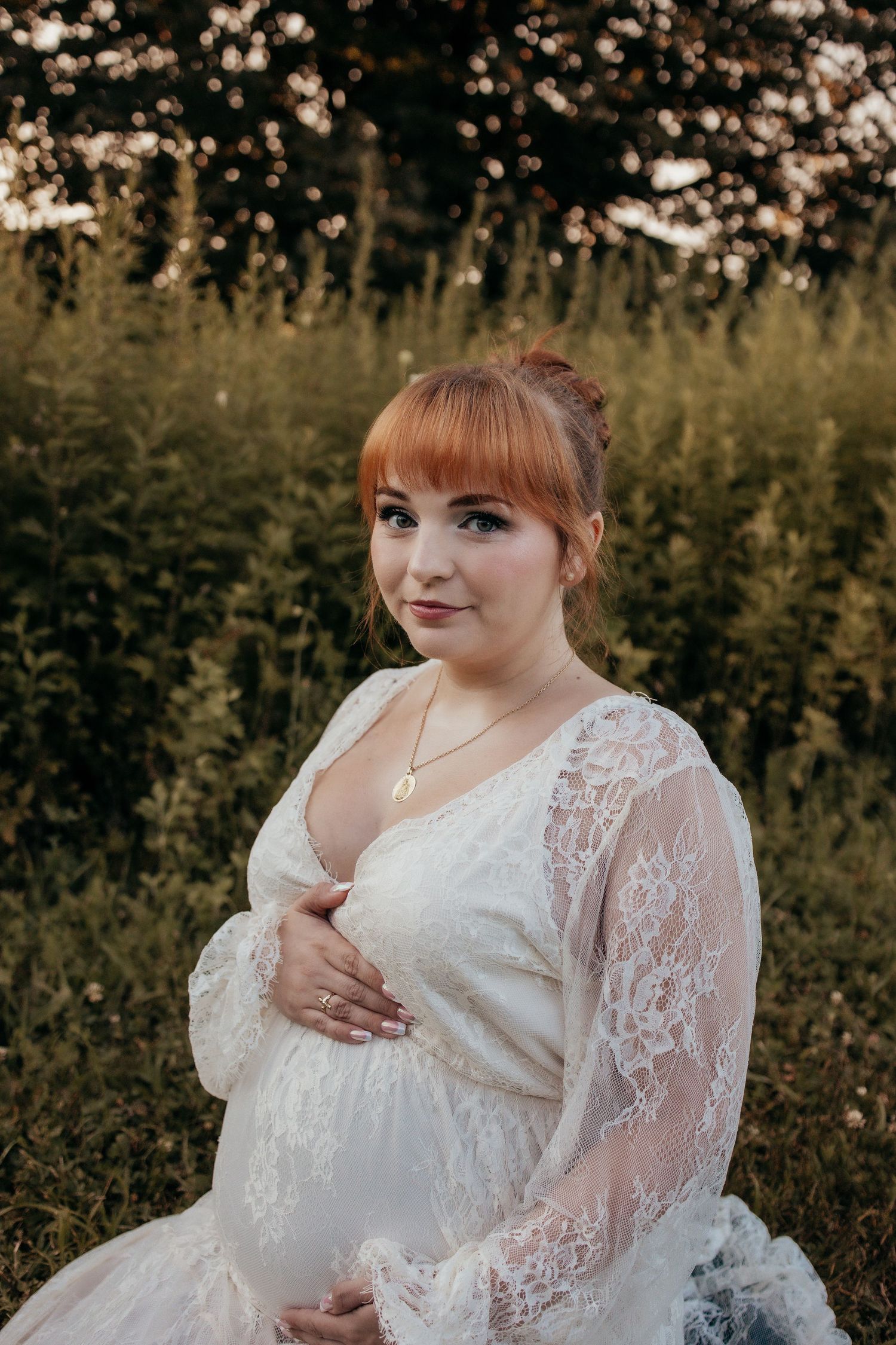 Pregnant woman in white lace dress with red hair poses outdoors in golden hour light among green foliage.