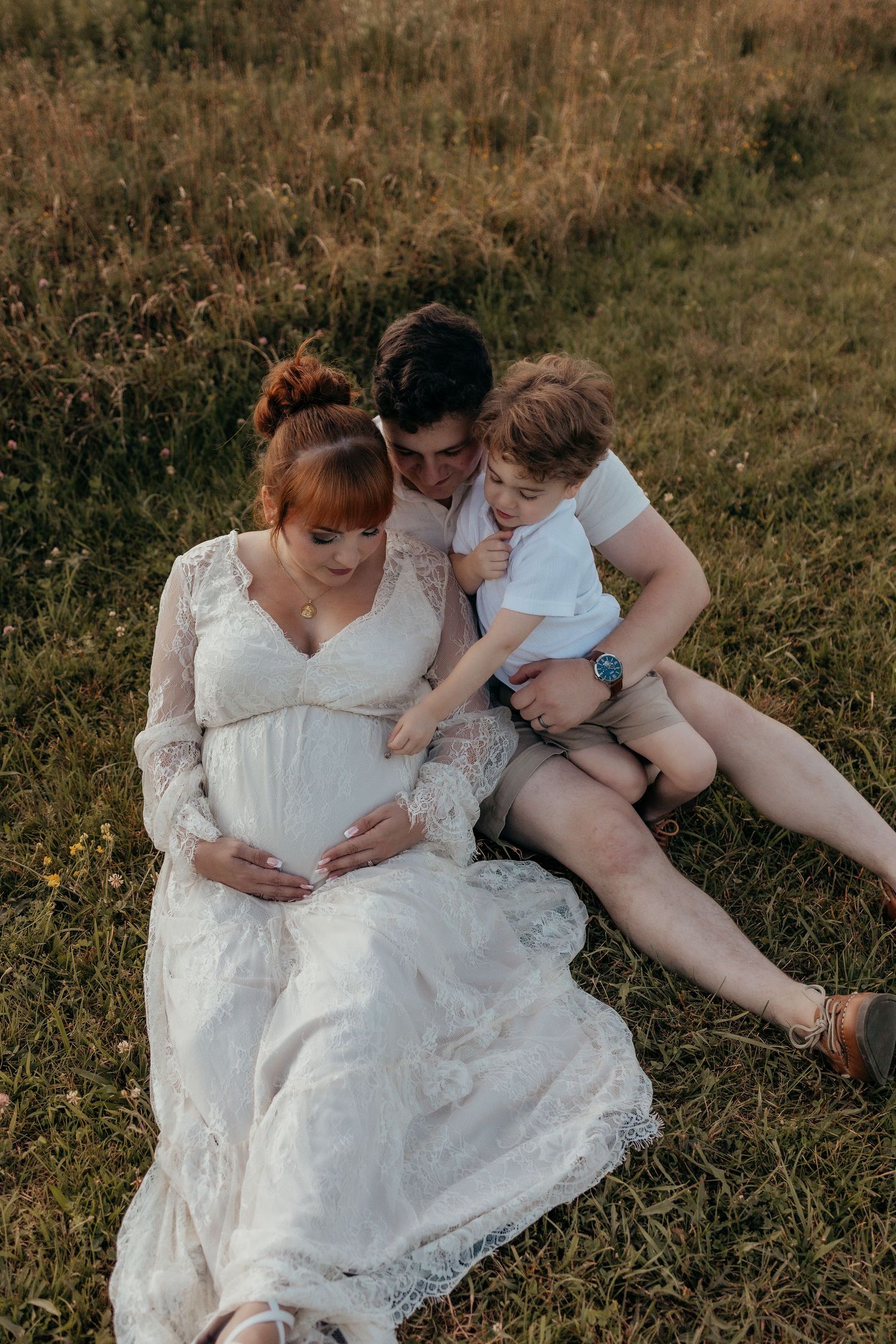 Pregnant woman in white lace dress sits with partner and young child in golden field during maternity photo session.