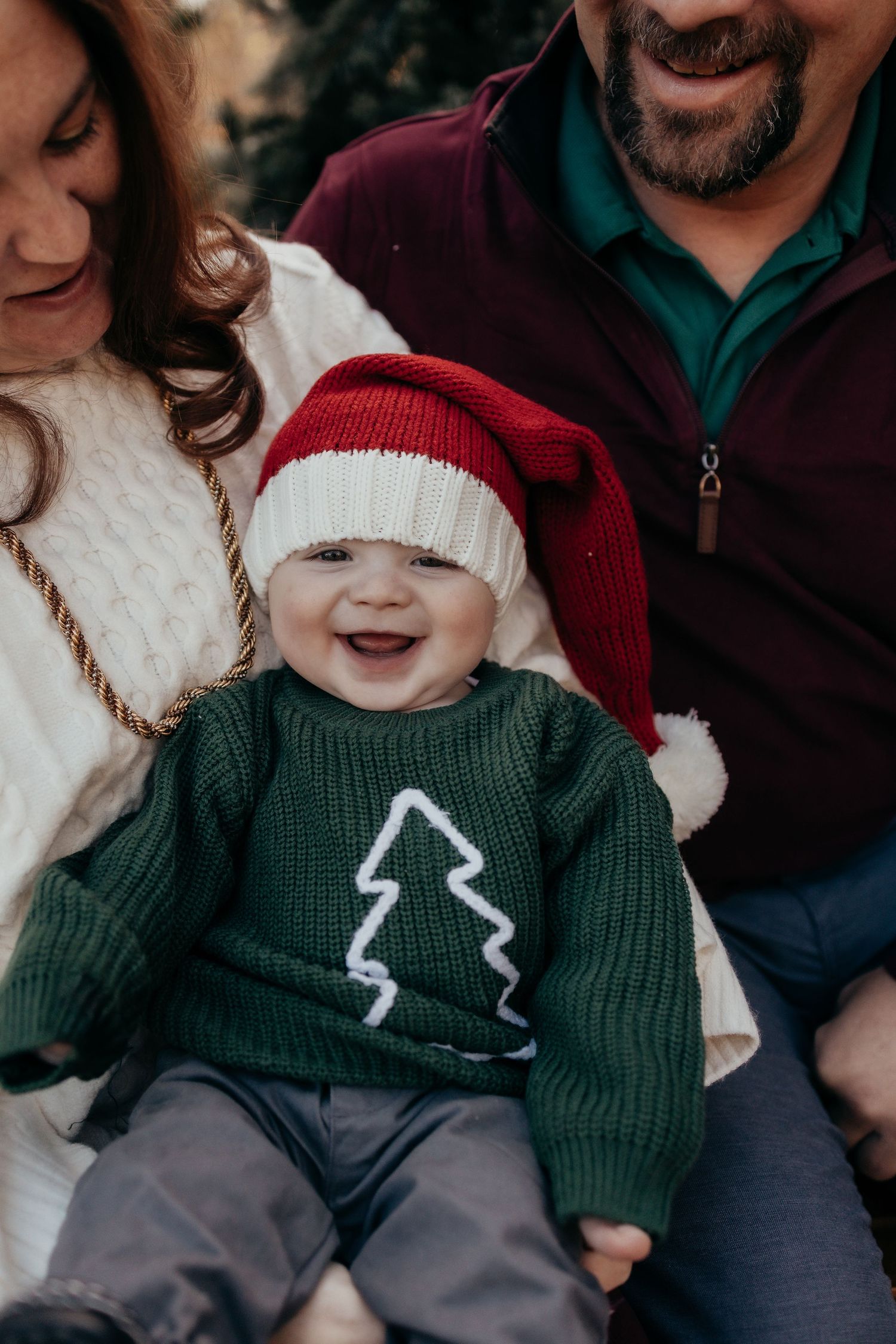 Smiling baby in Santa hat and green Christmas tree sweater sitting with parents during holiday family photo session.