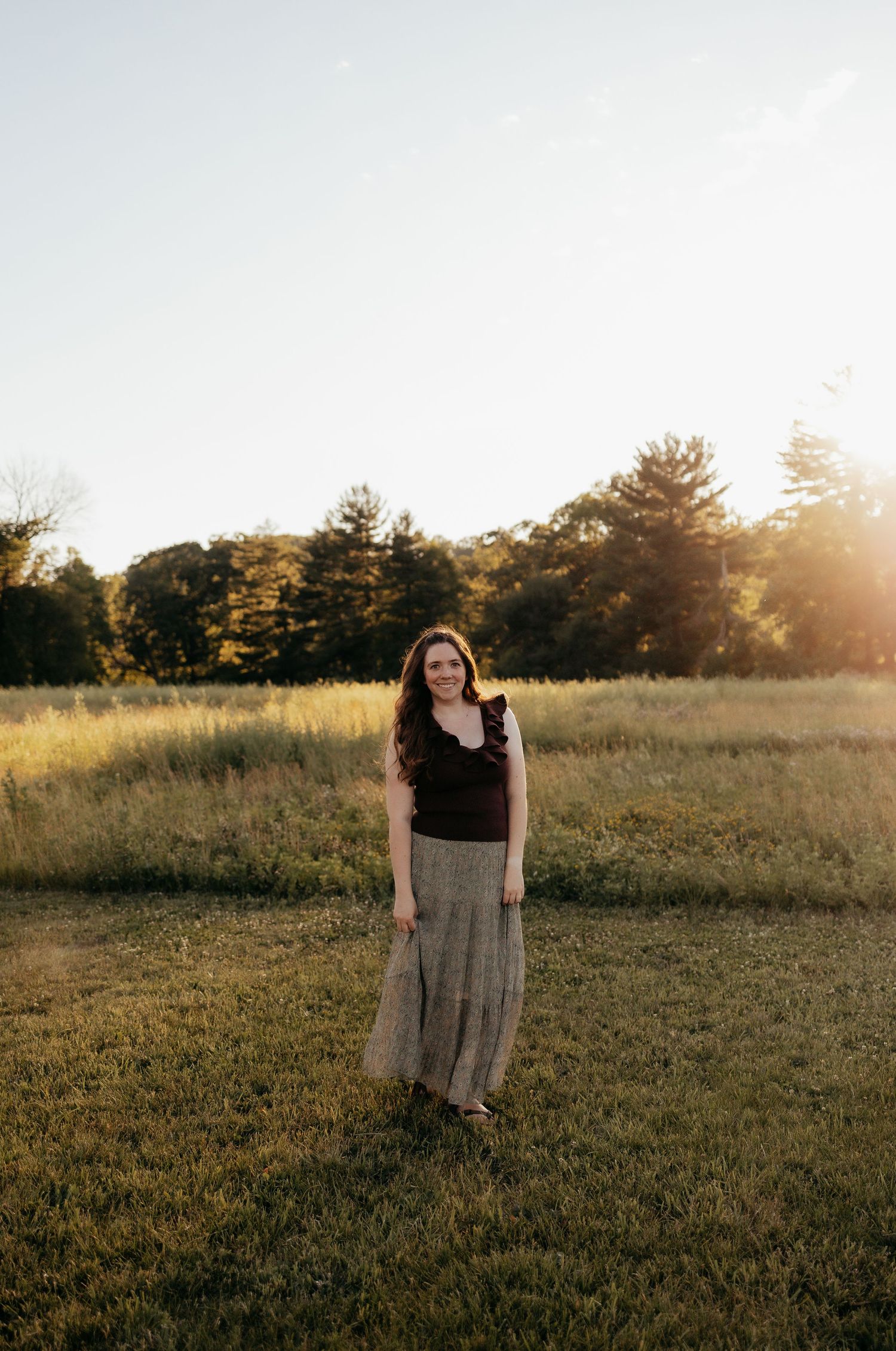 Young woman in black top and flowy skirt standing in sunlit meadow at golden hour.