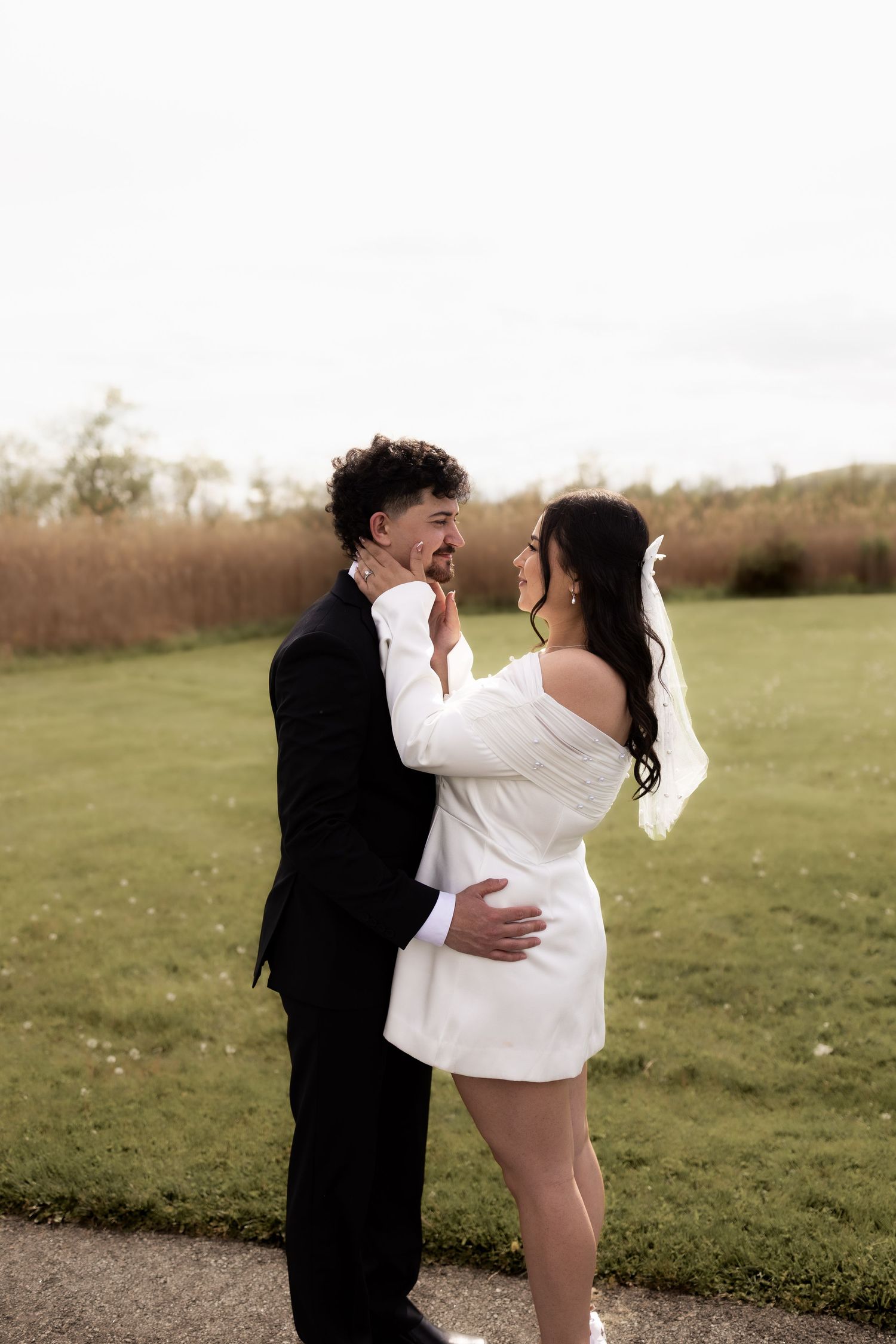 Bride and groom embracing and smiling at each other on a path surrounded by green fields.