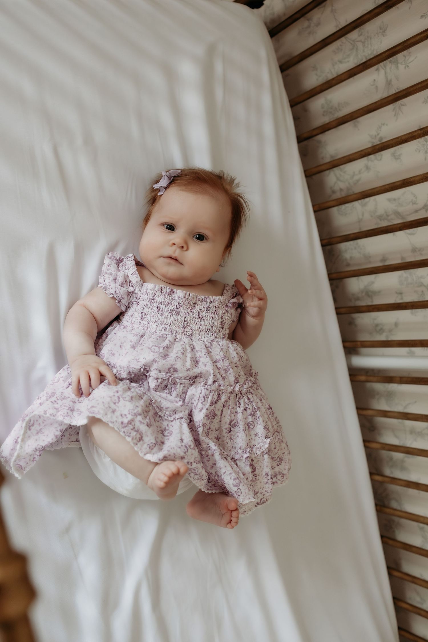Adorable baby girl in a floral pink dress lying in a white crib with wooden slats.
