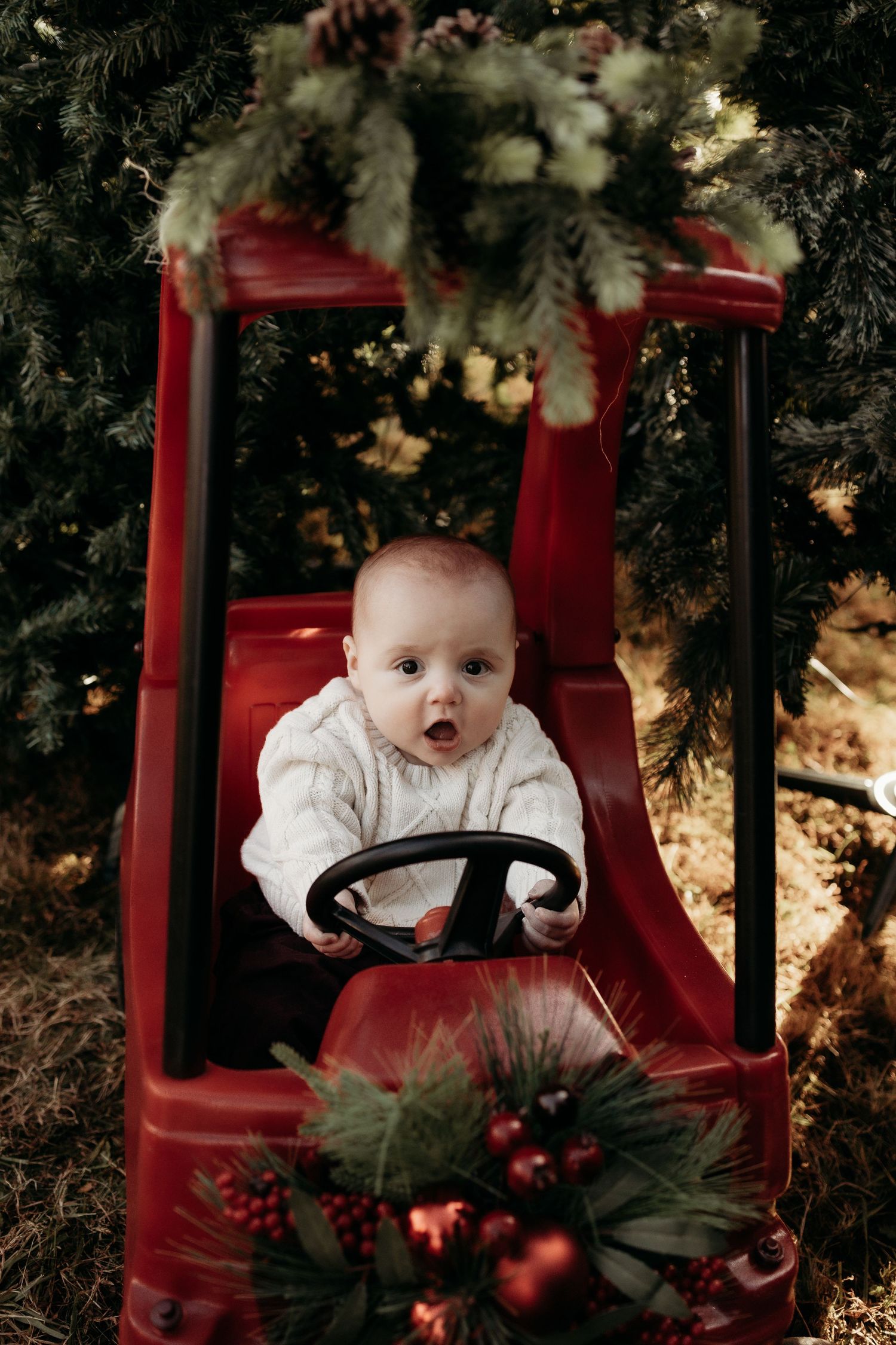Adorable baby in white sweater sitting in red toy truck surrounded by Christmas trees and holiday decorations.