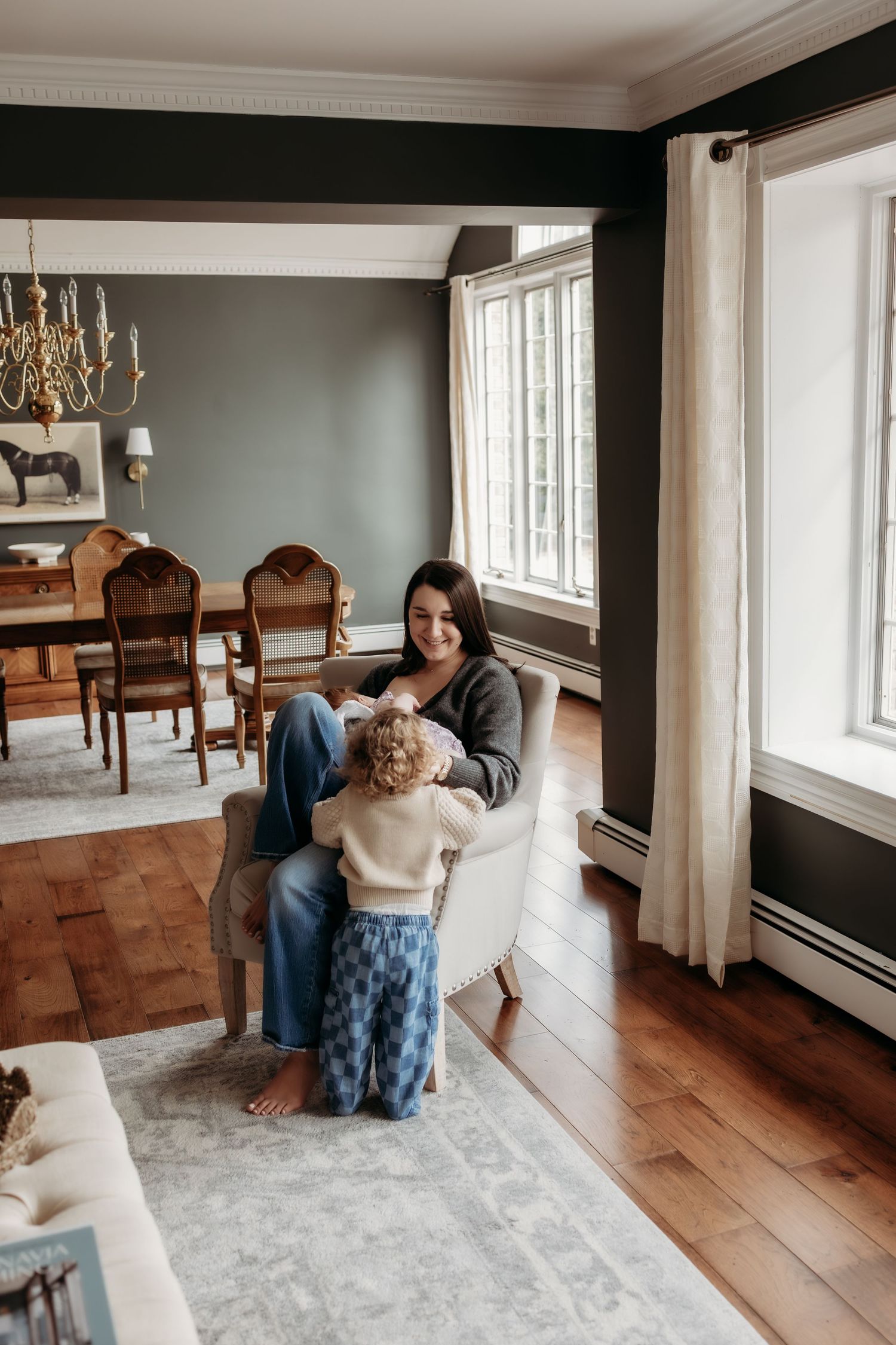 A woman sits in an armchair smiling at a toddler in a cozy home with hardwood floors and elegant dining room.