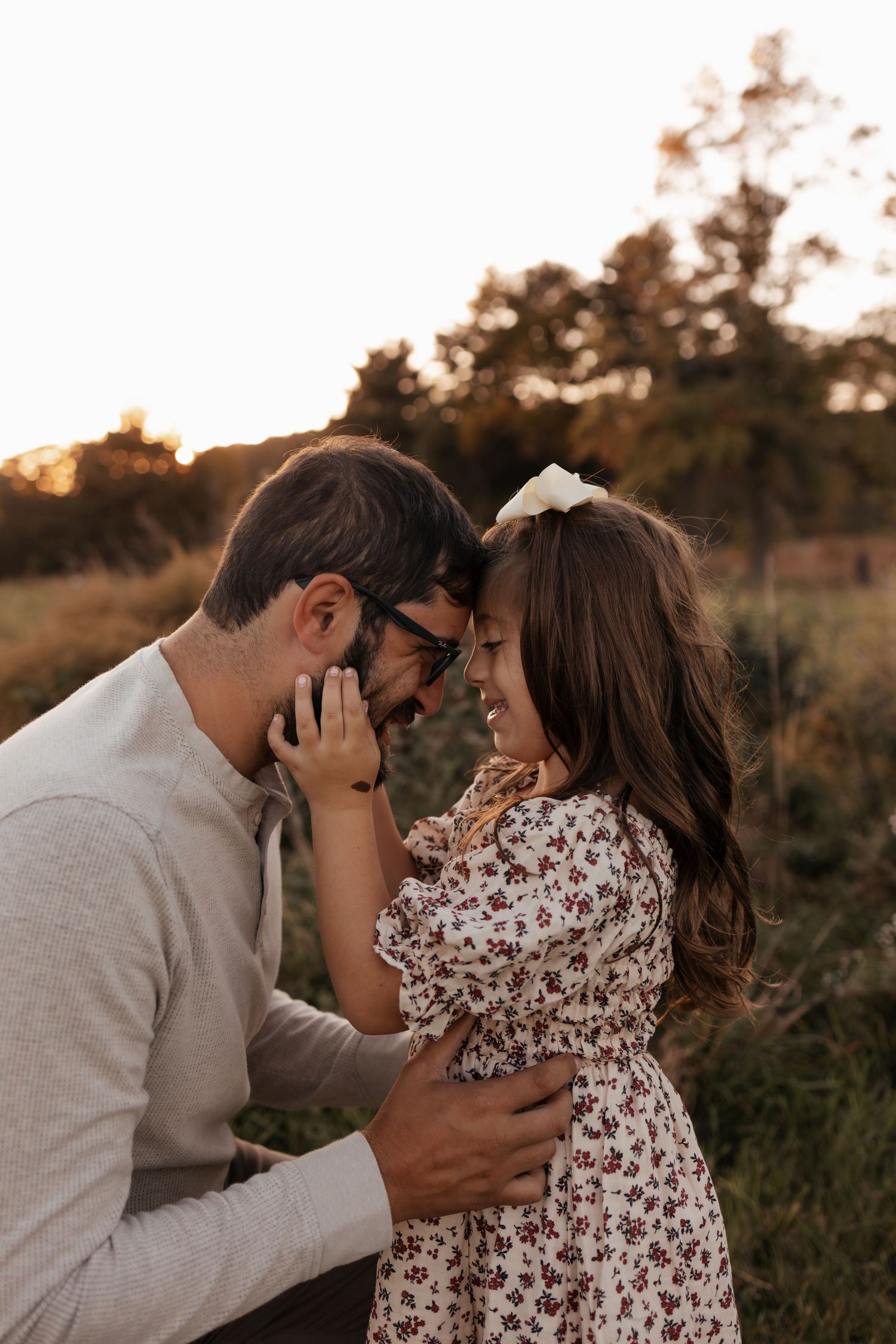 A bearded man and a young girl in a floral dress touch foreheads and smile in a golden-lit field at sunset.