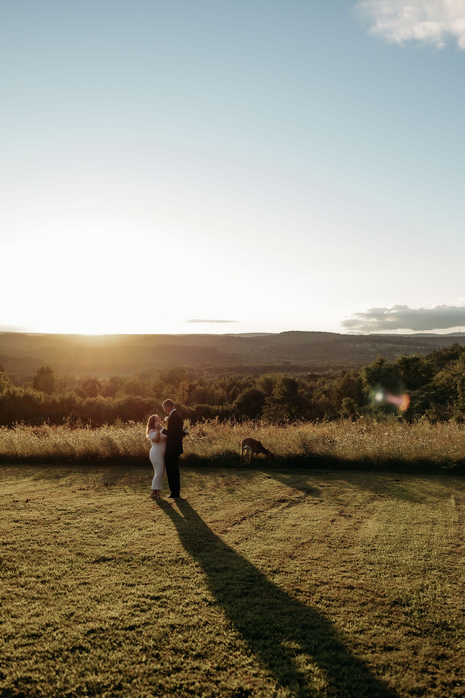 Couple embracing at golden hour sunset in a meadow with rolling hills and a dog nearby.