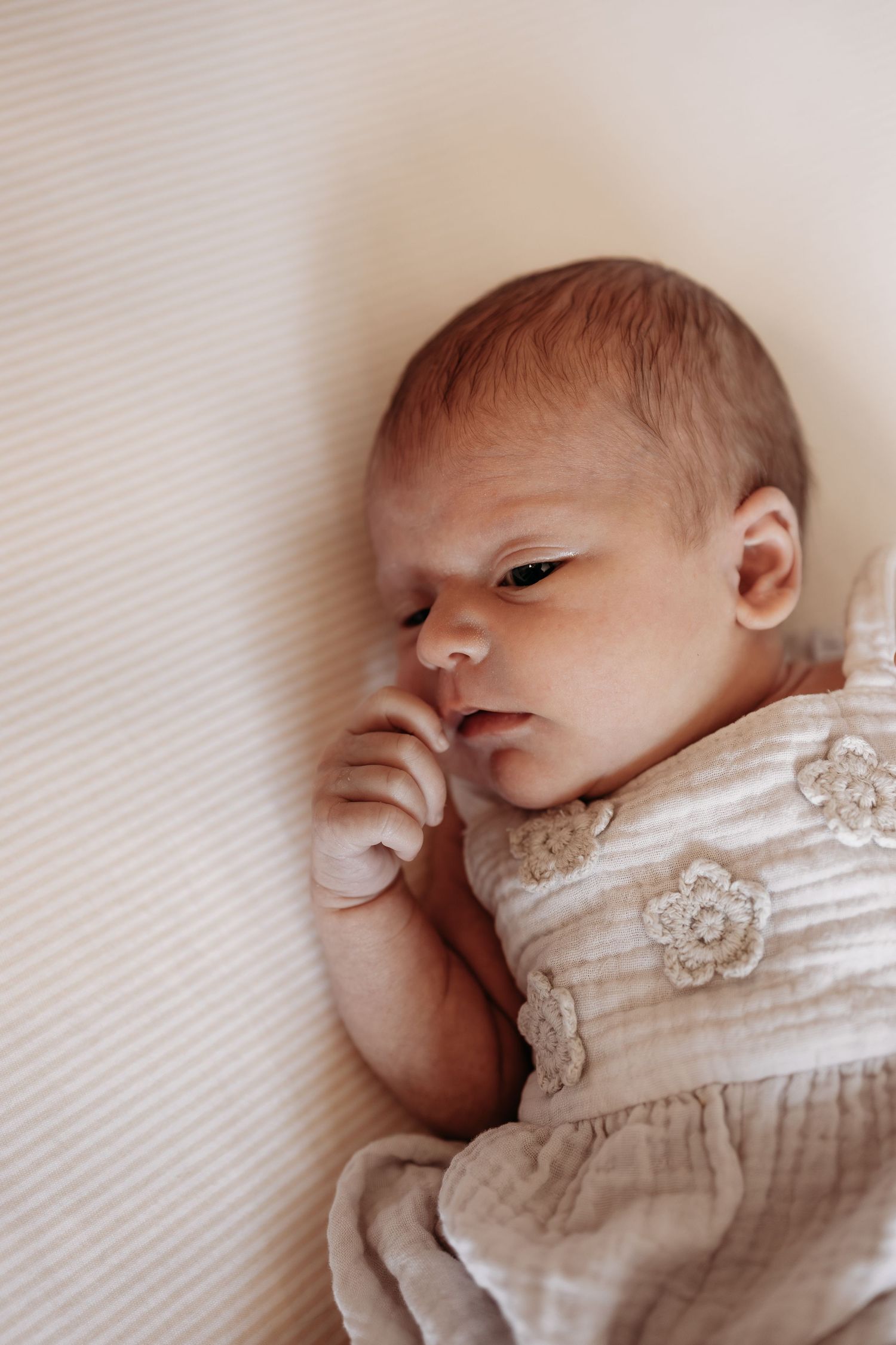 Newborn baby lying on white sheet wearing floral crochet romper with tiny hand raised to face.