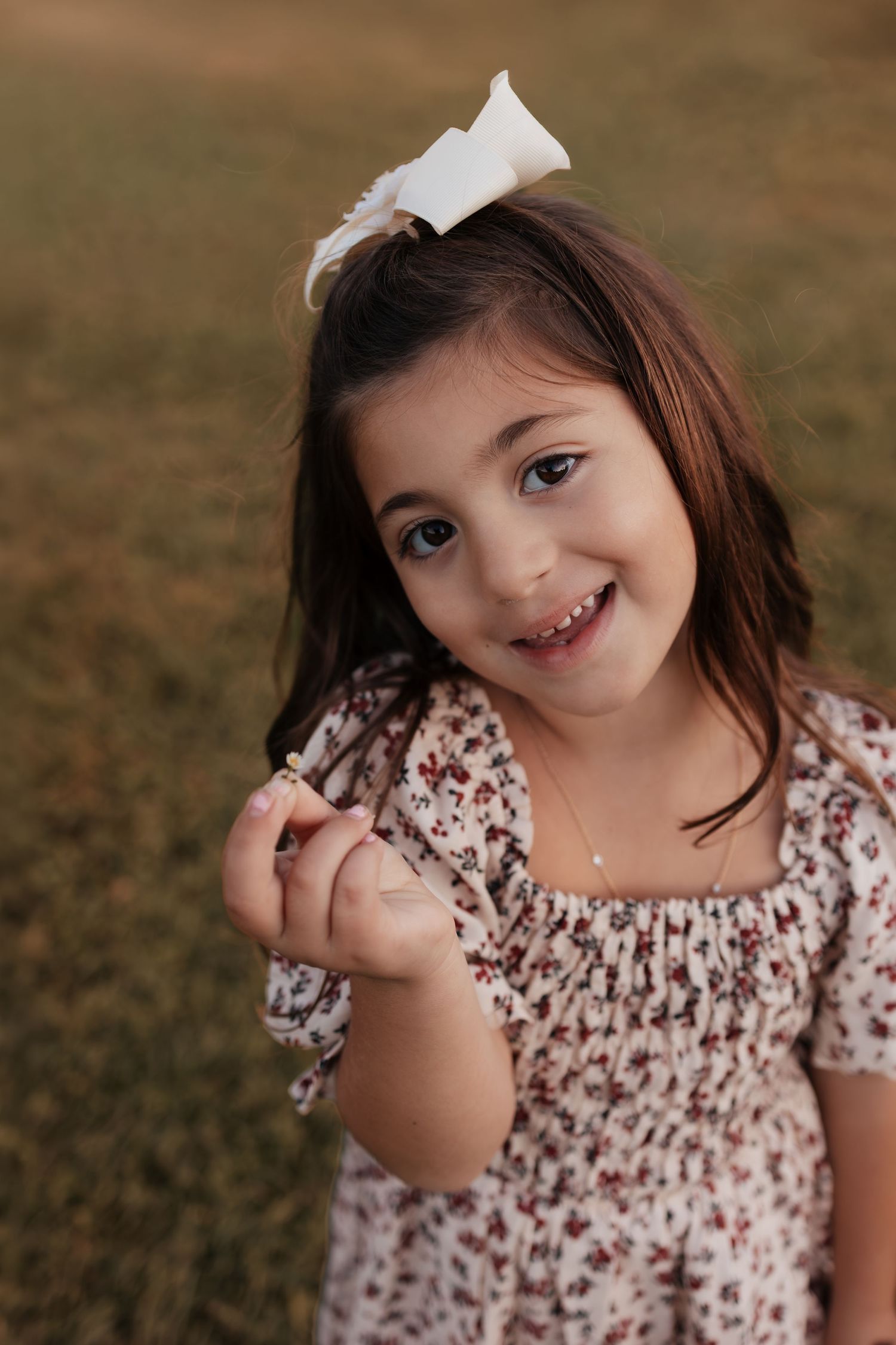 Smiling young girl with a white hair bow and floral smocked dress holding a small flower while standing on a grassy field.