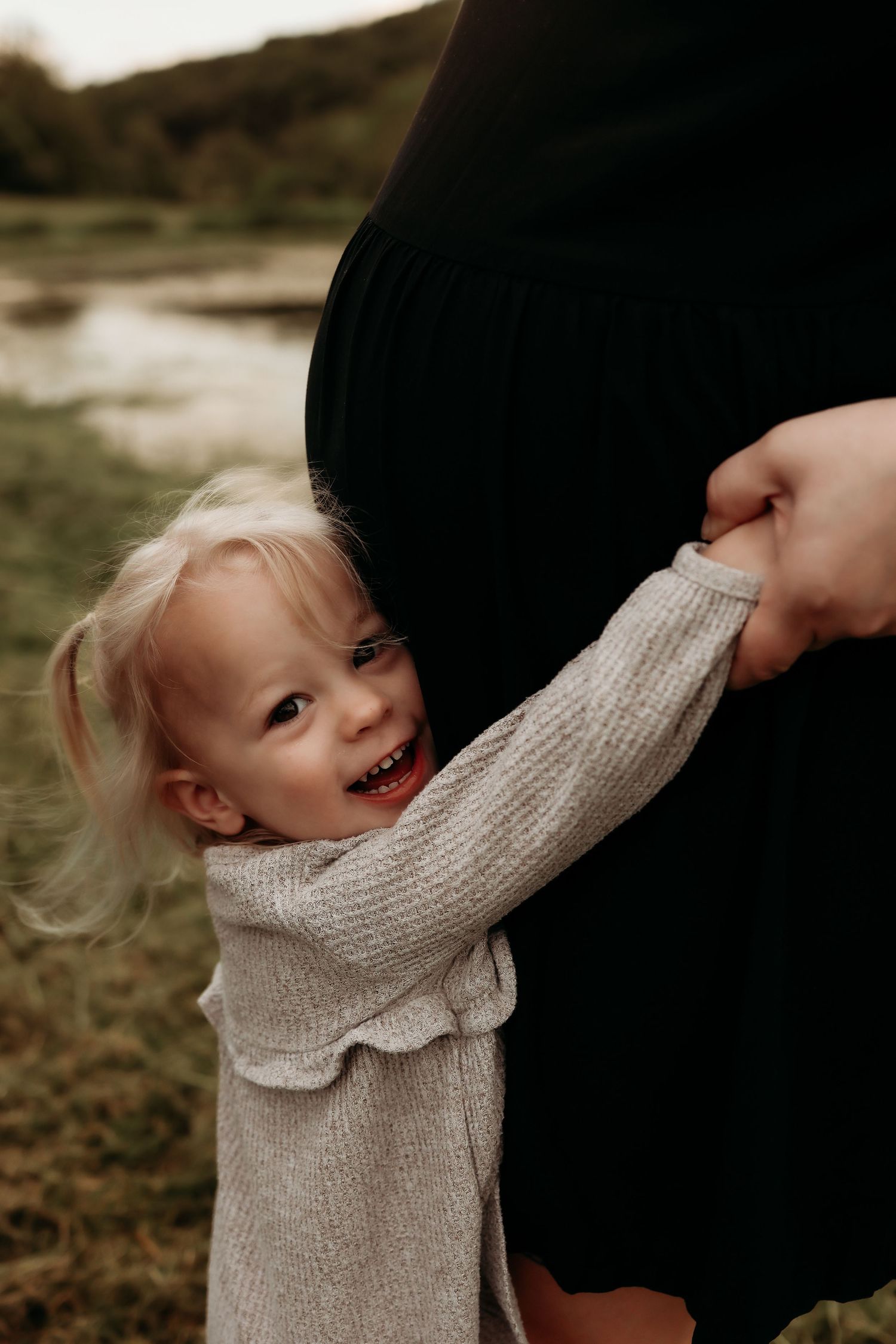 A happy toddler girl hugs her pregnant mothers baby bump in an outdoor setting.