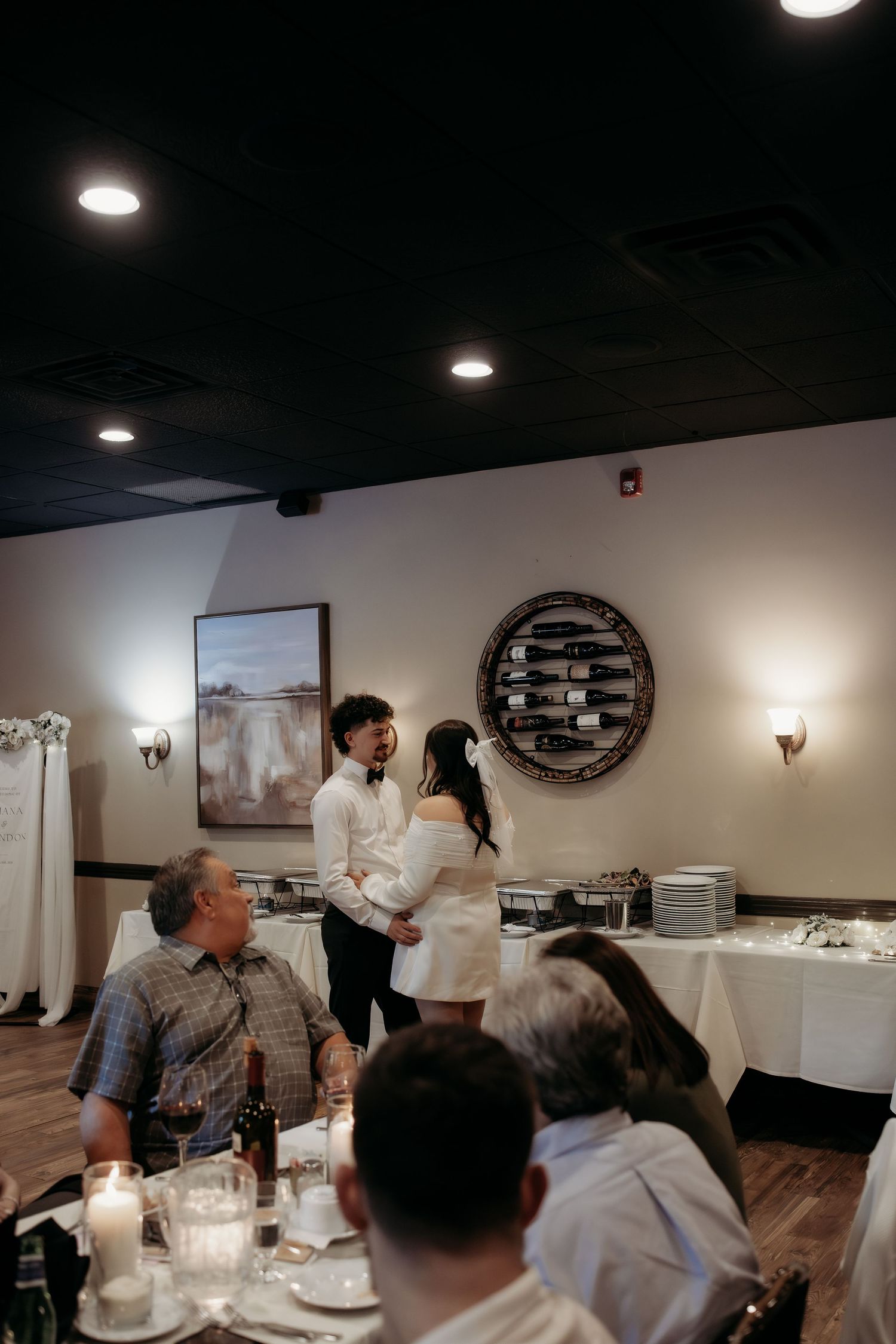 Couple sharing joyful moment during wedding reception in dimly lit restaurant with wine rack.