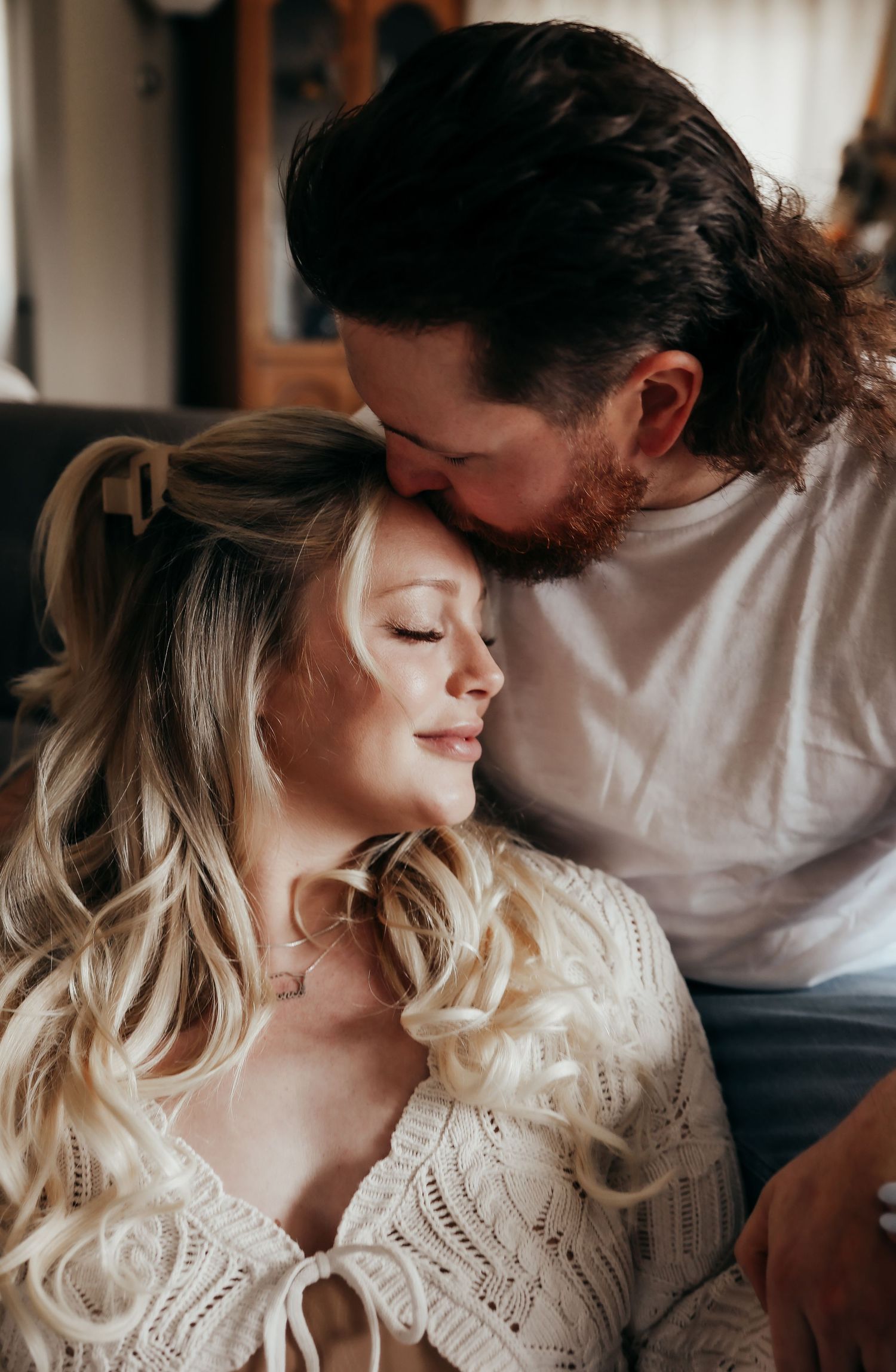 Bearded man gently kissing blonde woman's head in an intimate maternity moment.