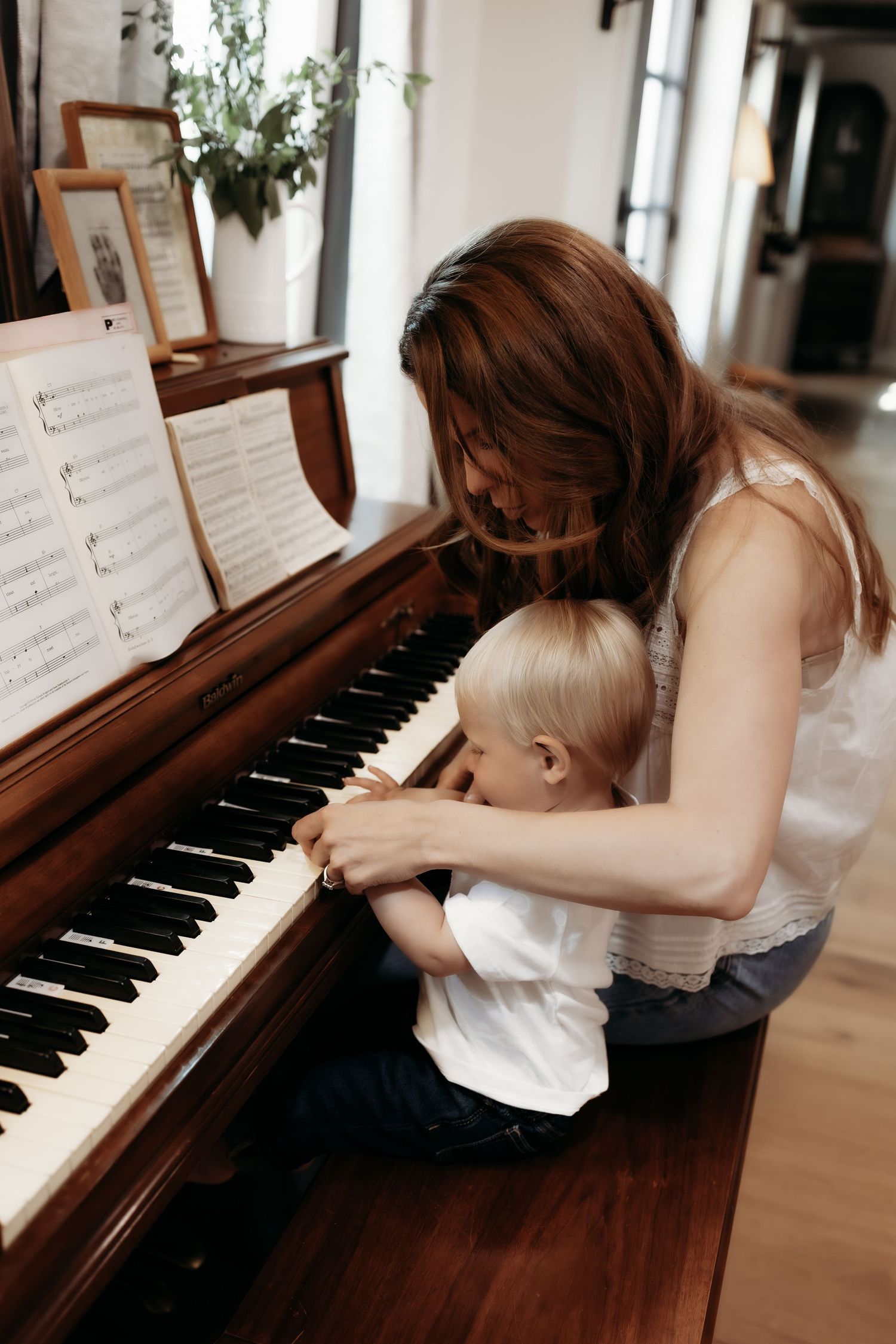 Mother teaching young toddler to play piano on wooden upright piano indoors.