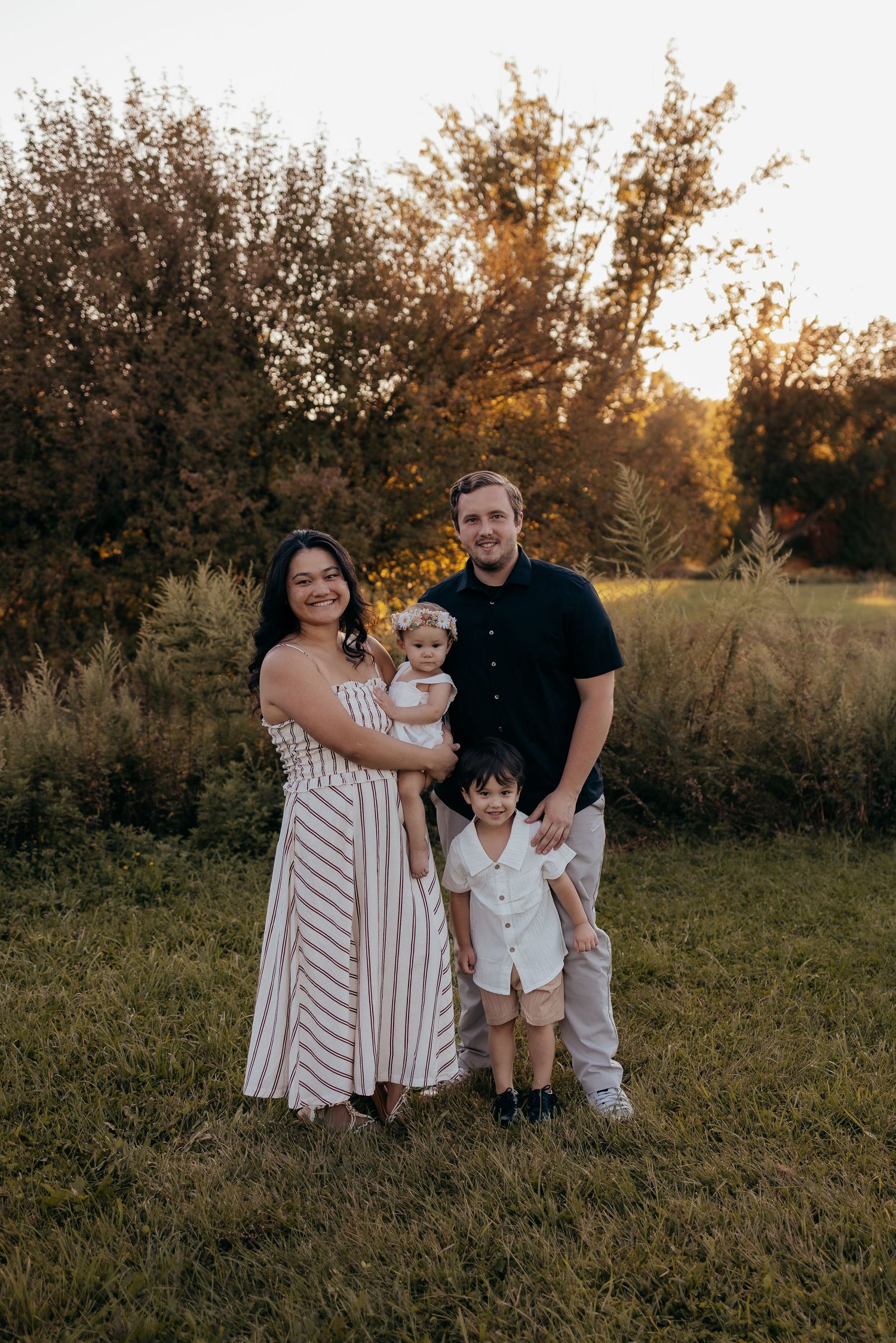 Family of four posing outdoors at golden hour, mom in striped dress, dad in navy shirt, with toddler son and baby daughter.