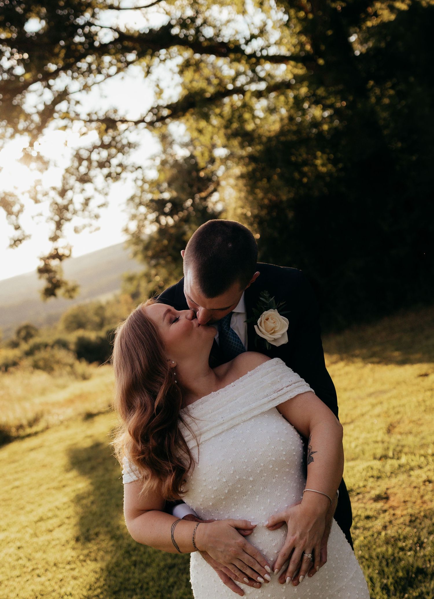 A bride and groom share a romantic kiss outdoors at golden hour, surrounded by lush green trees and rolling hills.