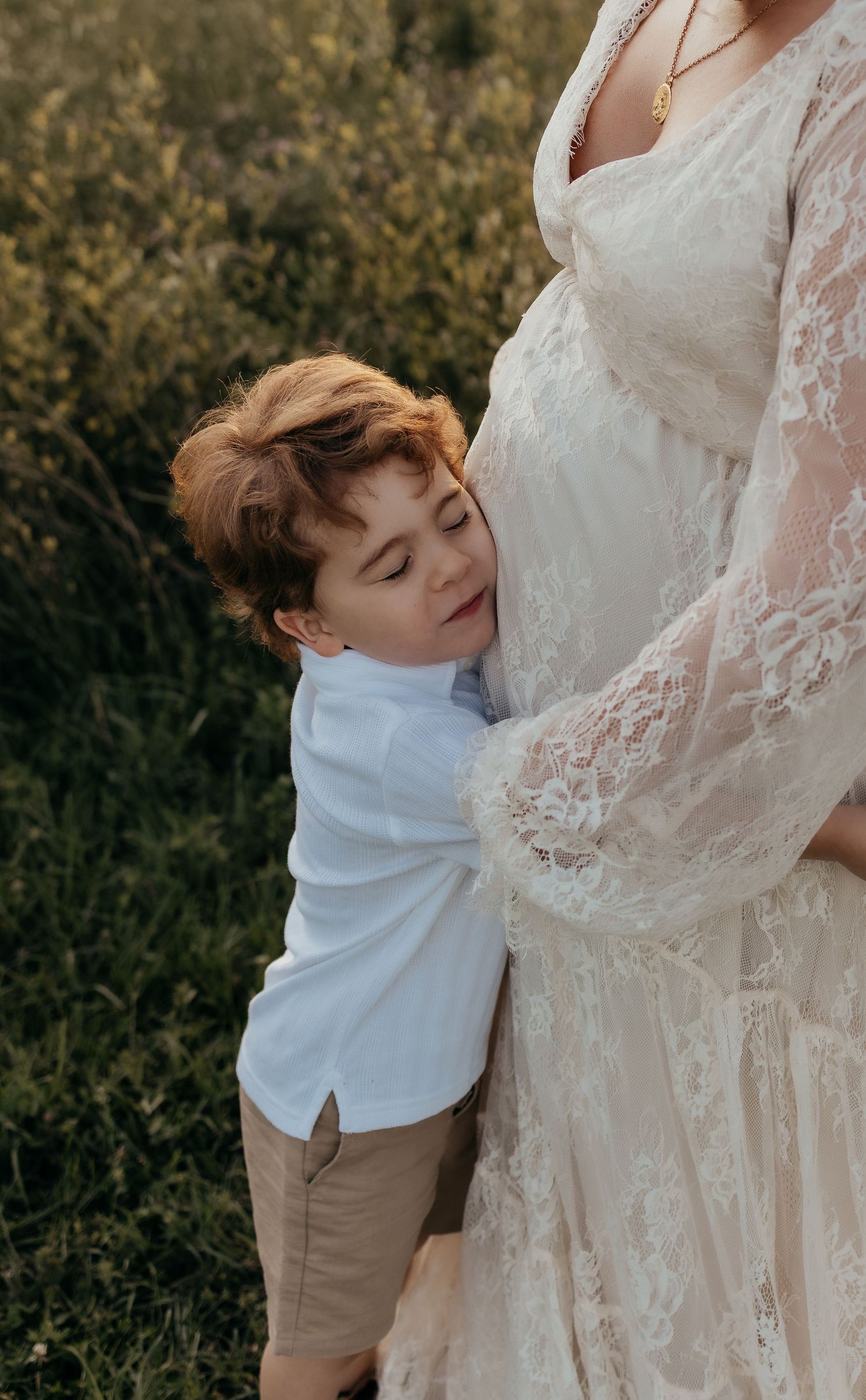 A young boy hugs his pregnant mother's belly outdoors, both dressed in white and khaki tones.