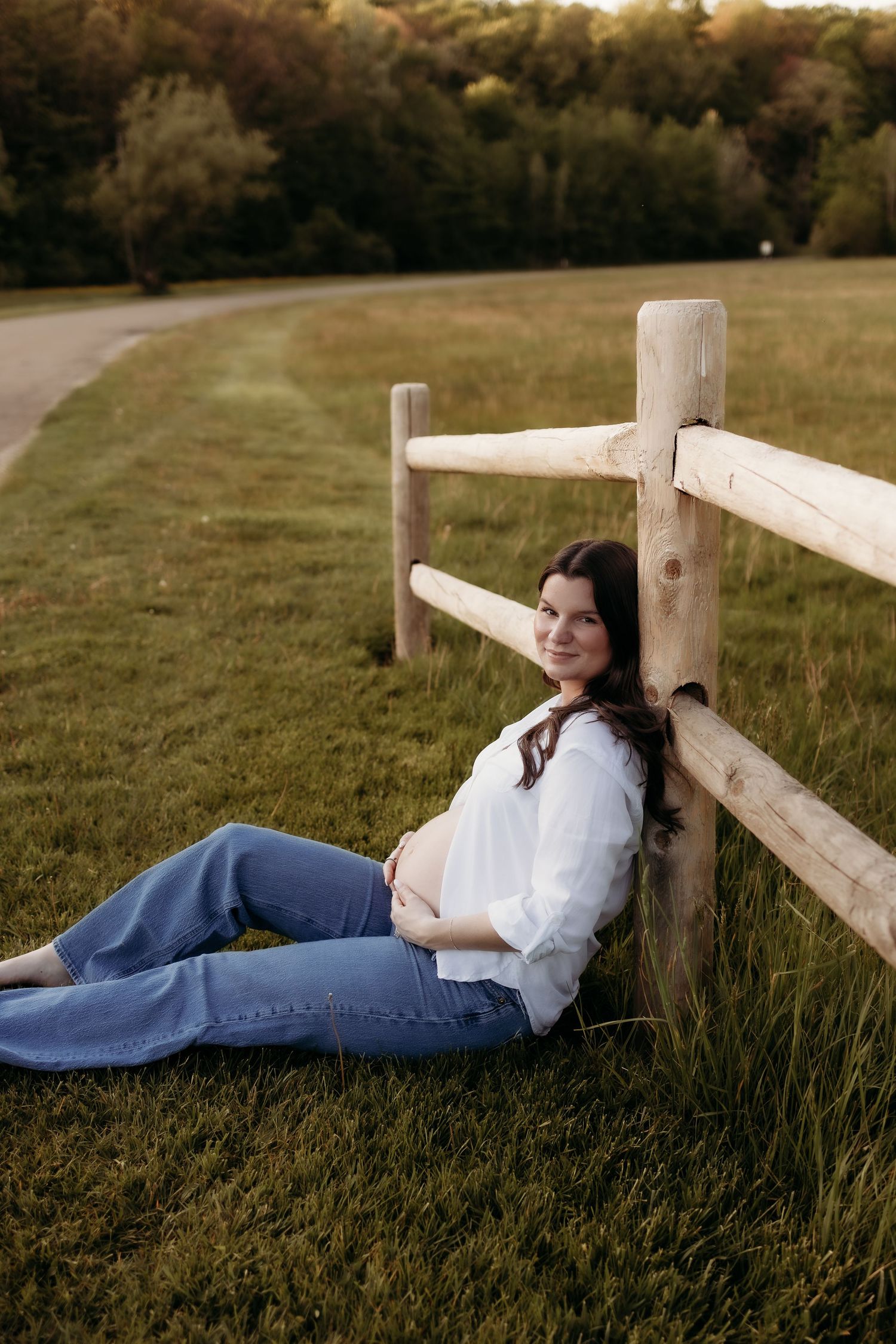 Pregnant woman in white shirt and jeans sitting against wooden fence in a lush green field at sunset.