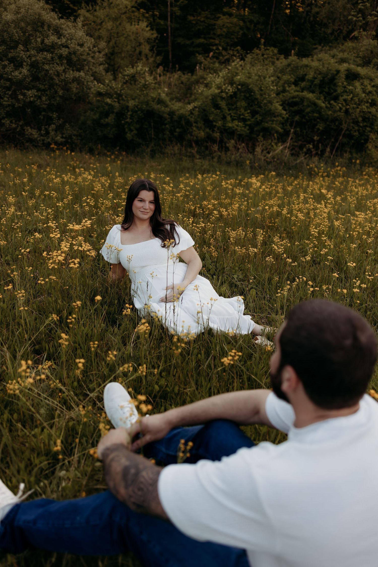 Pregnant woman in white dress sits smiling in yellow wildflower field while partner relaxes nearby.