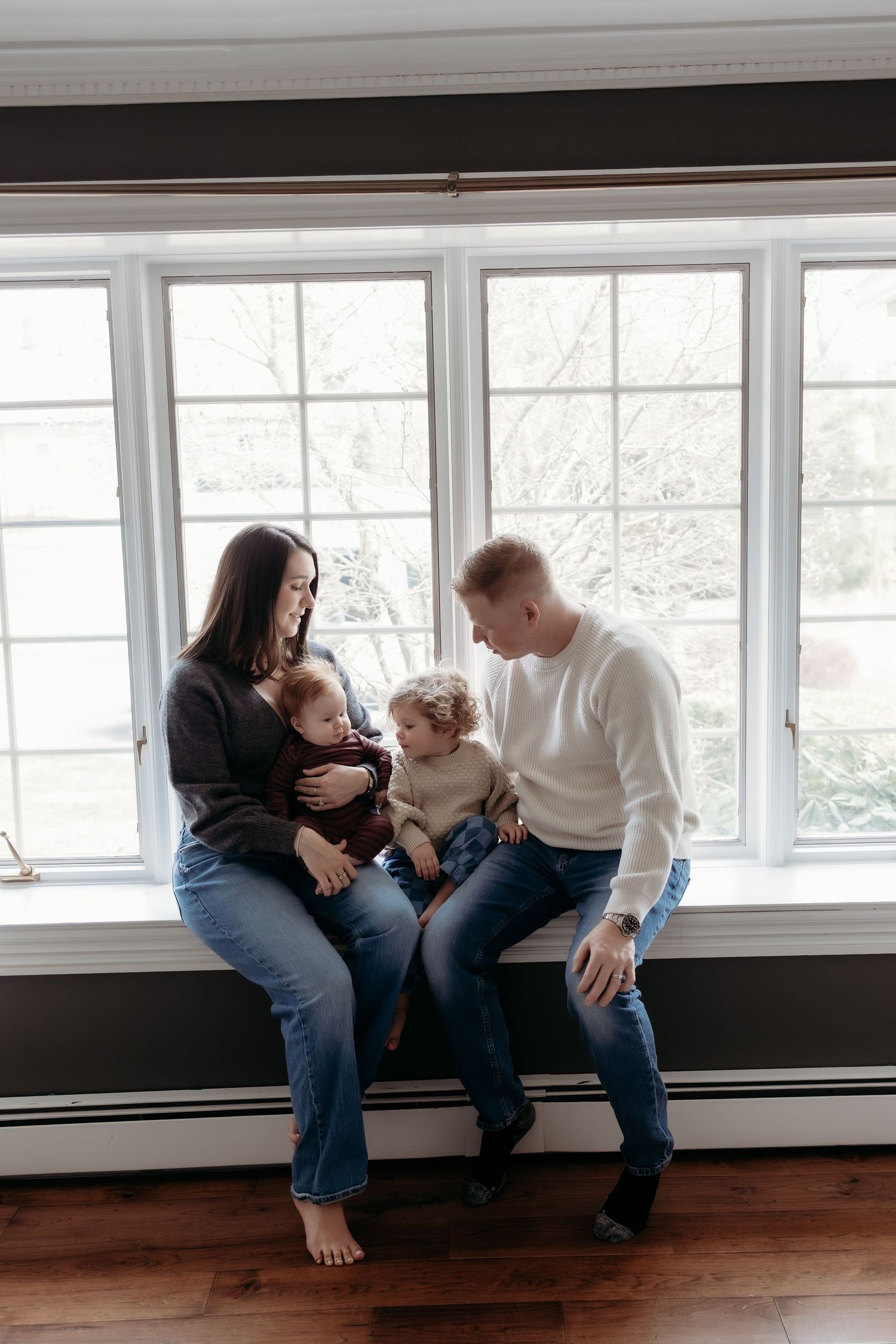 A young family of four sits together on a window ledge, sharing a warm intimate moment.