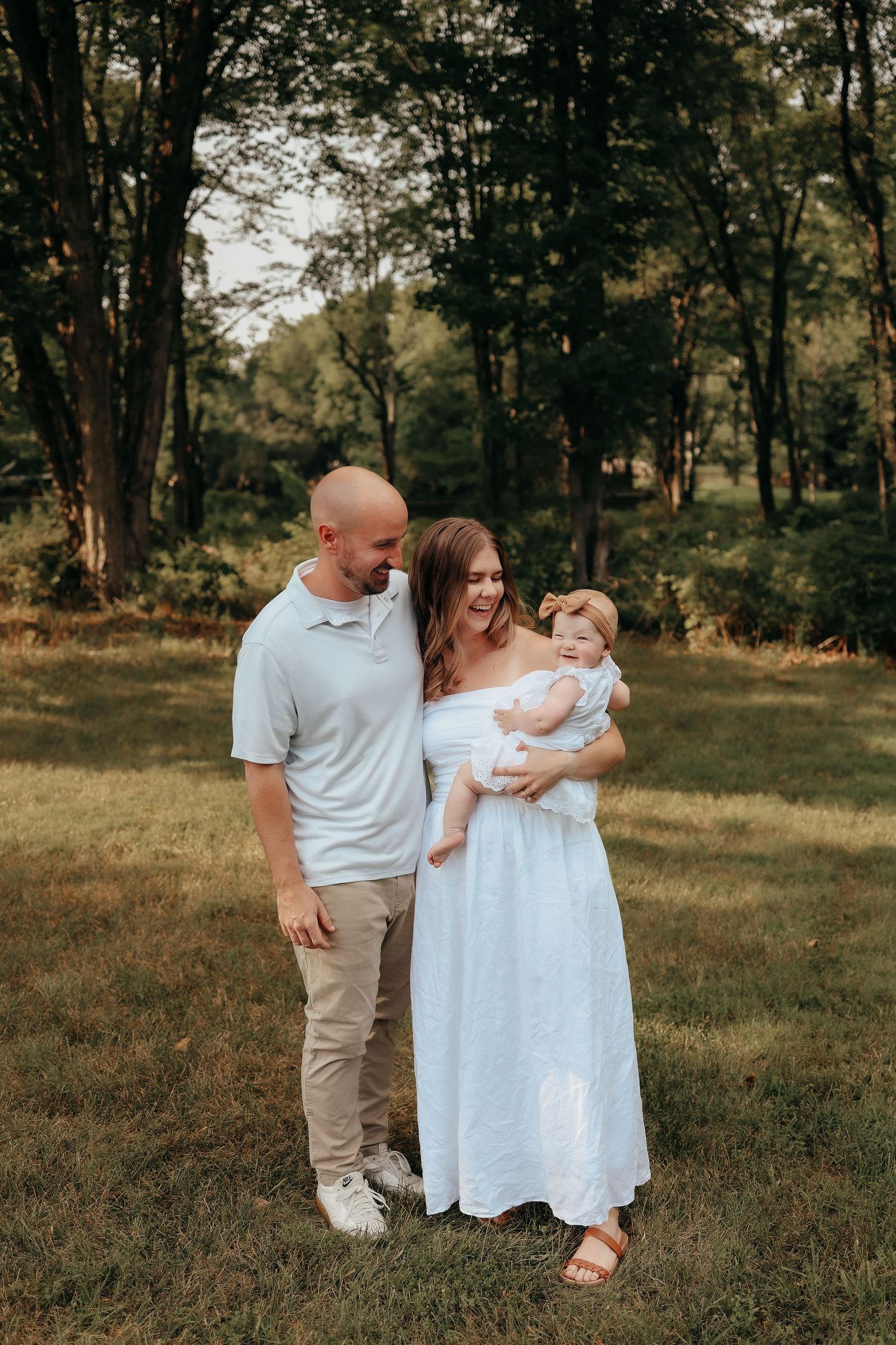 Happy family portrait with bald father, mother in blue dress holding infant in wooded park.