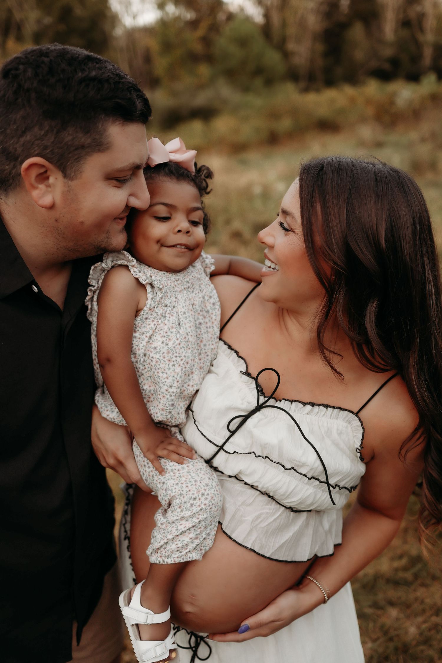Expecting parents smiling at toddler daughter in floral outfit during outdoor maternity photo session.