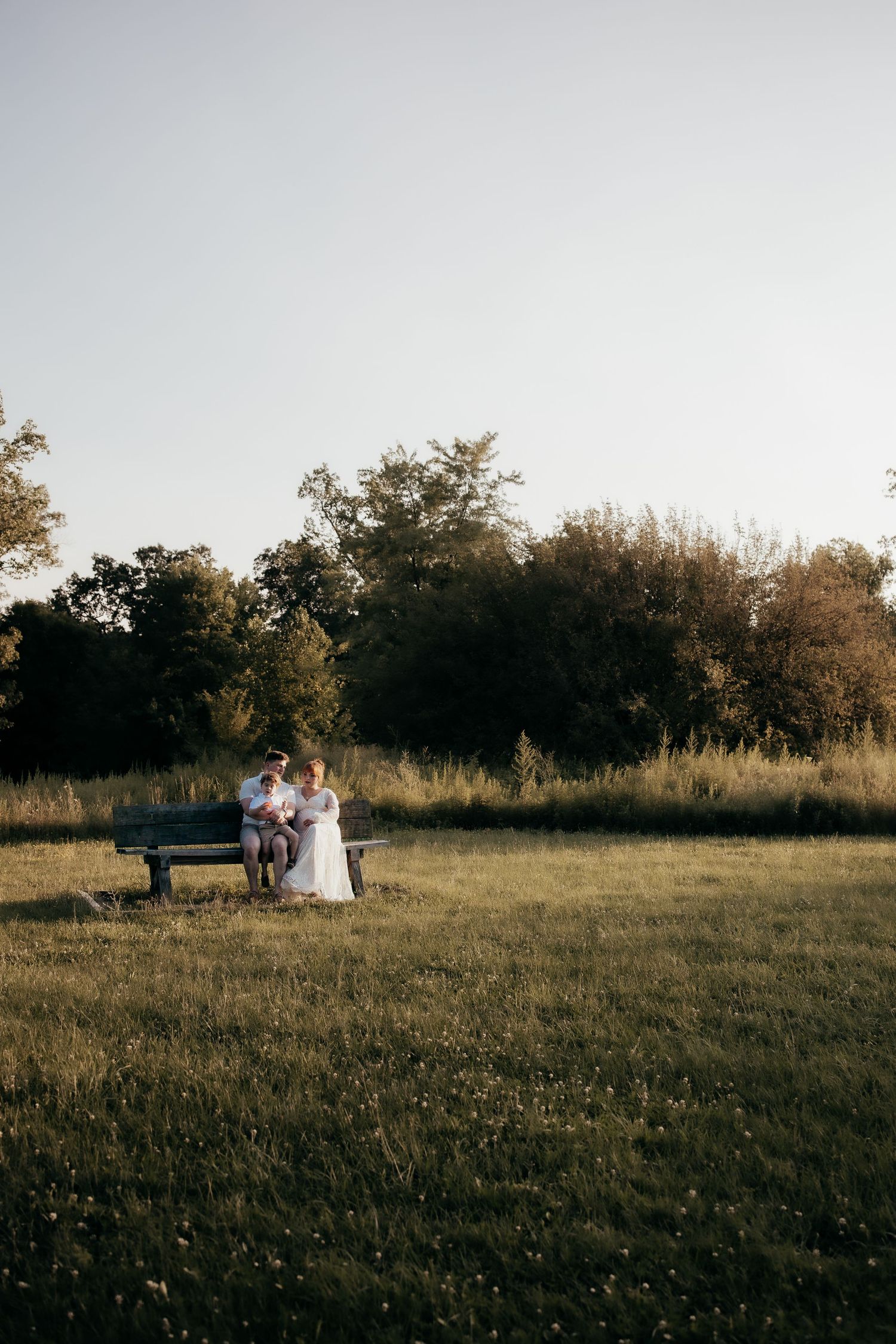 Couple in wedding attire sitting on a bench in a sunlit green meadow surrounded by trees.