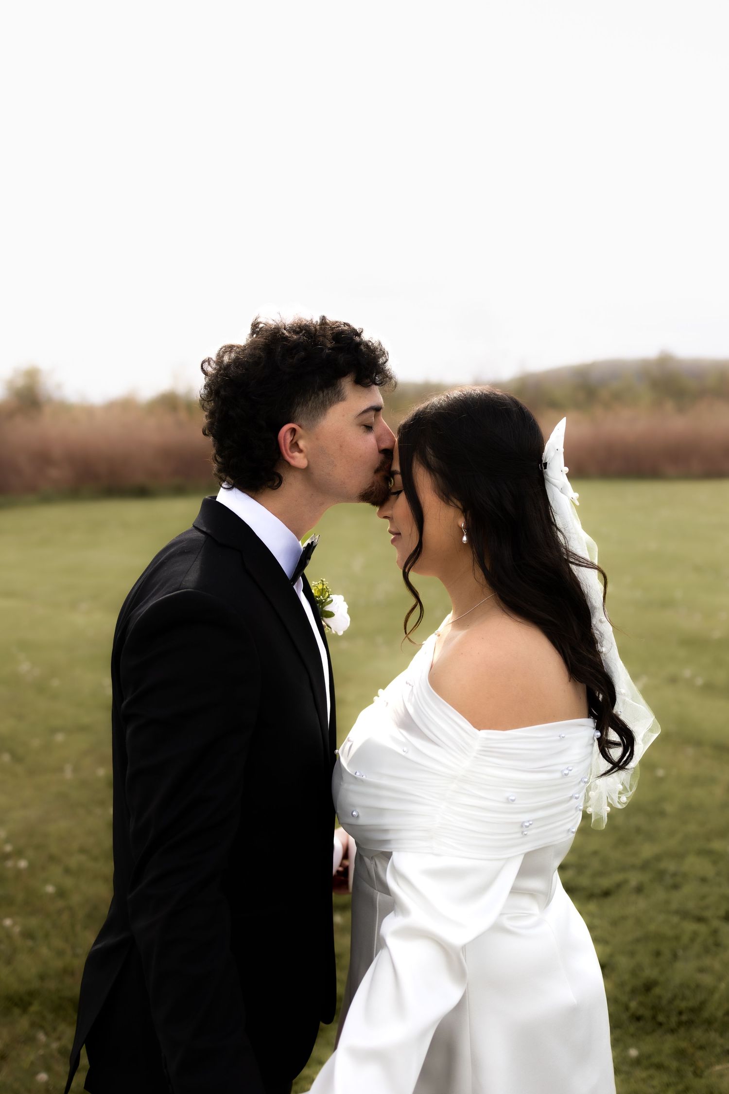 A groom in a black tuxedo leans in to kiss his bride in an off-shoulder white gown in a green field.