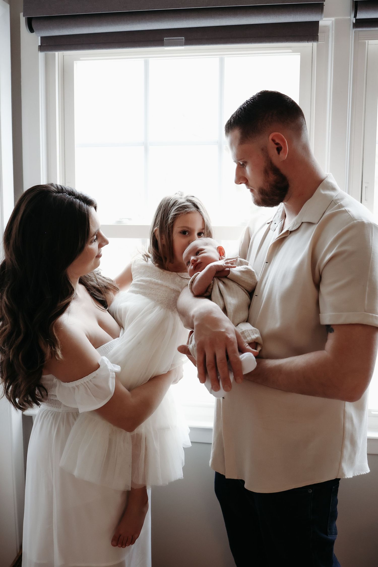 Family of four with newborn and toddler girl standing by bright window in soft natural light.