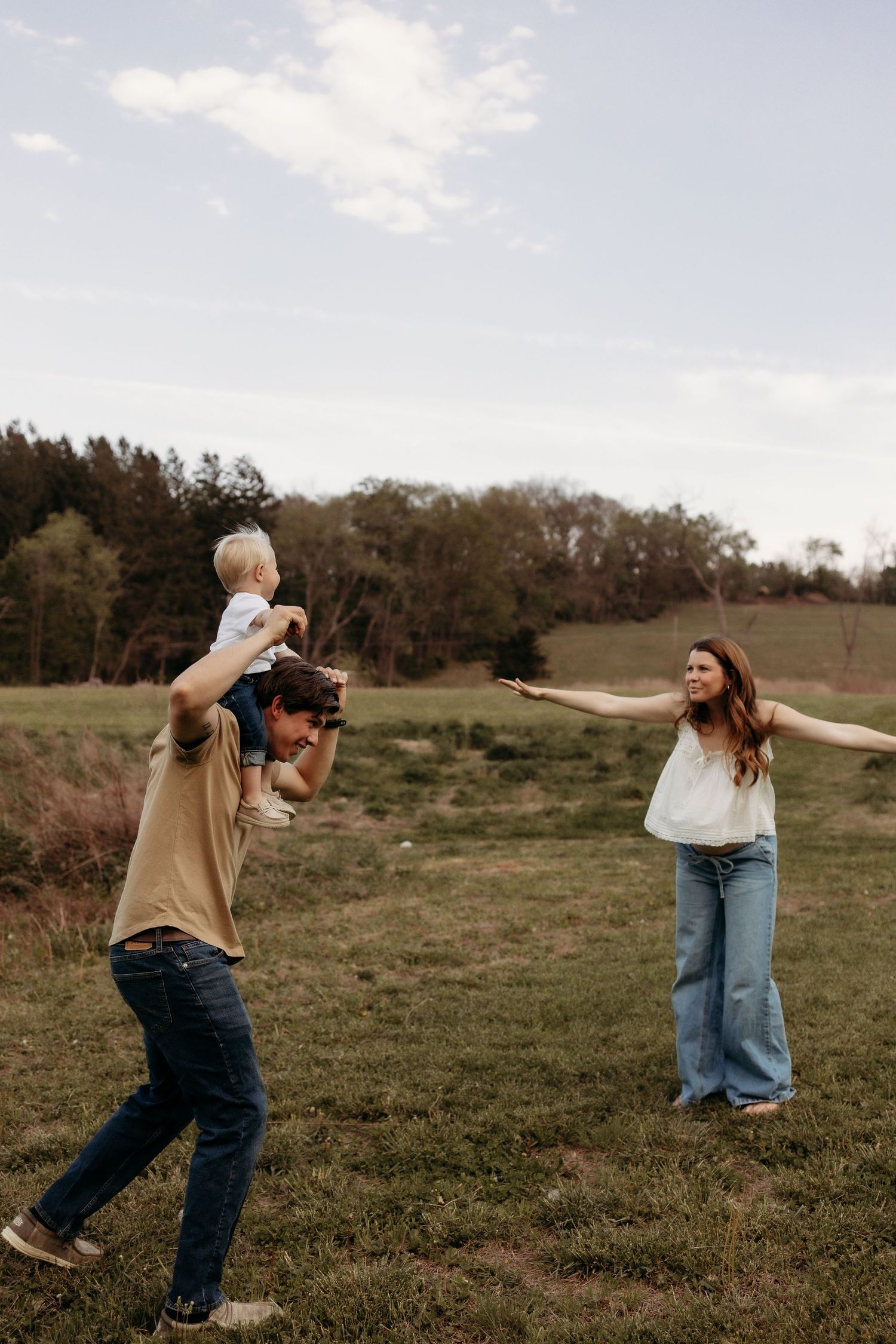 Family playing outdoors in a green field, father carrying toddler on shoulders while mother reaches out smiling.