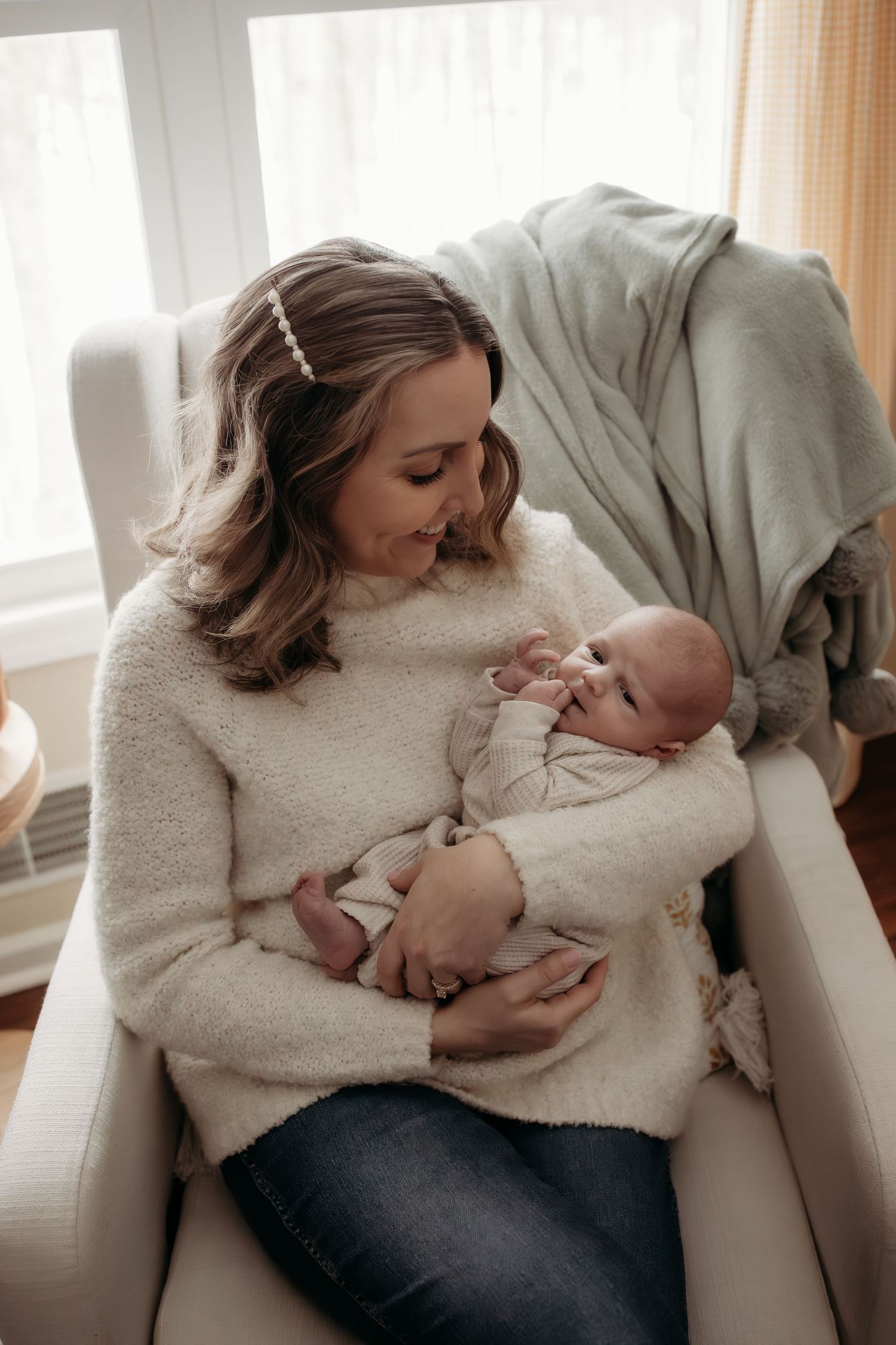 Mother in cream sweater gazes lovingly at newborn baby while seated in white nursery glider chair.