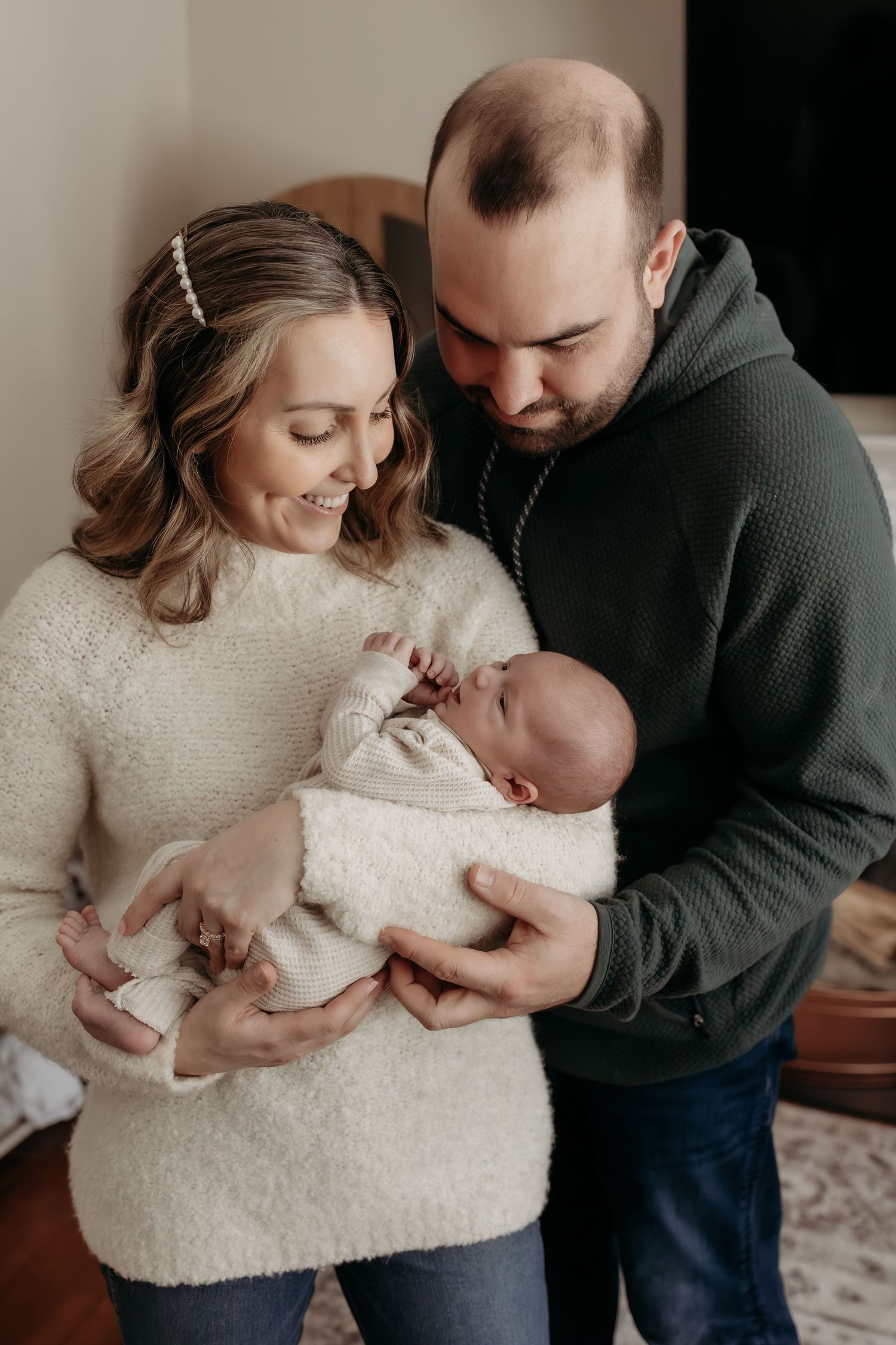 Parents in coordinating green and cream outfits gazing down at swaddled newborn baby together.