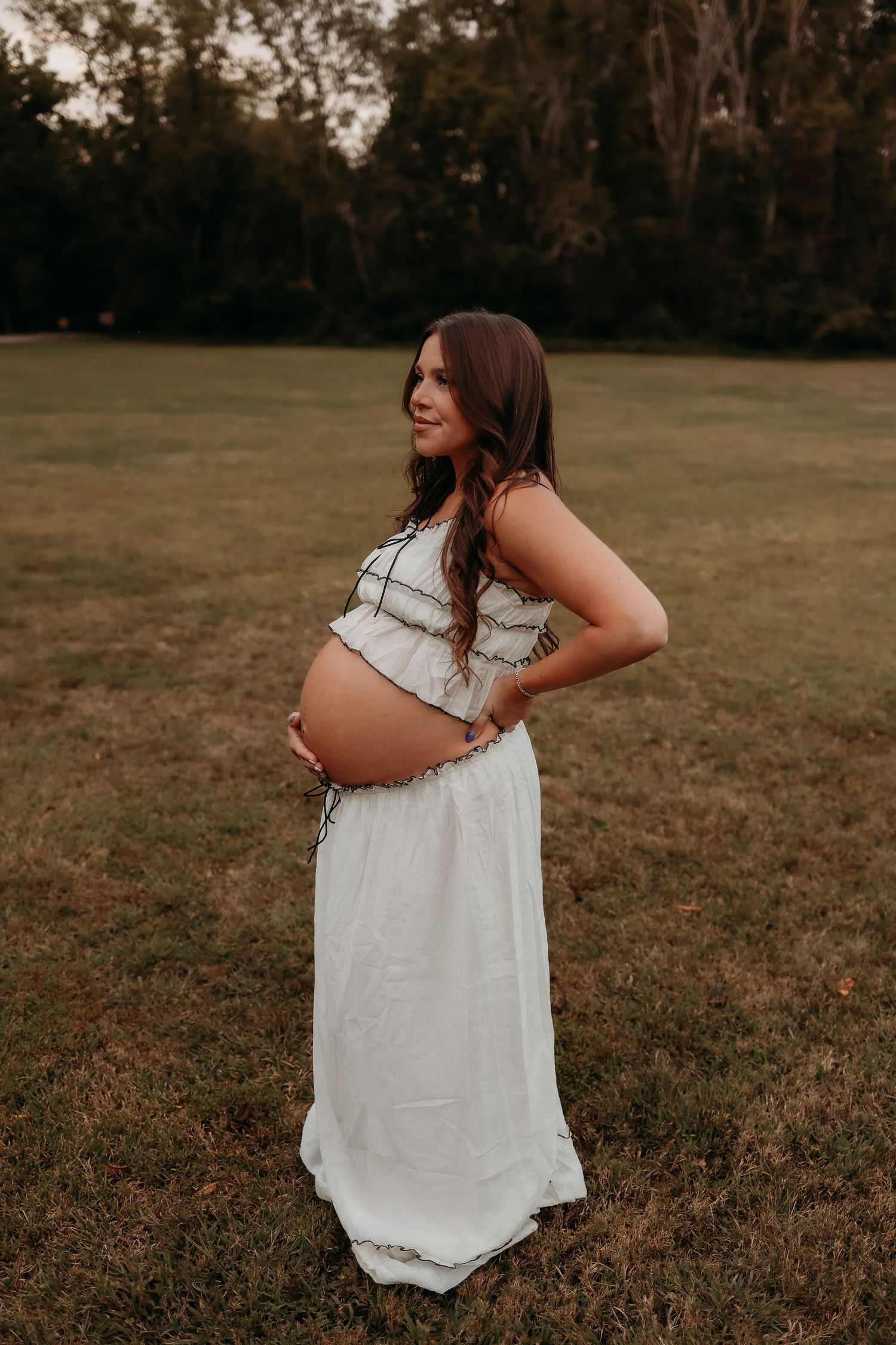 Pregnant woman in white dress posing sideways in an open field, showcasing her baby bump at sunset.
