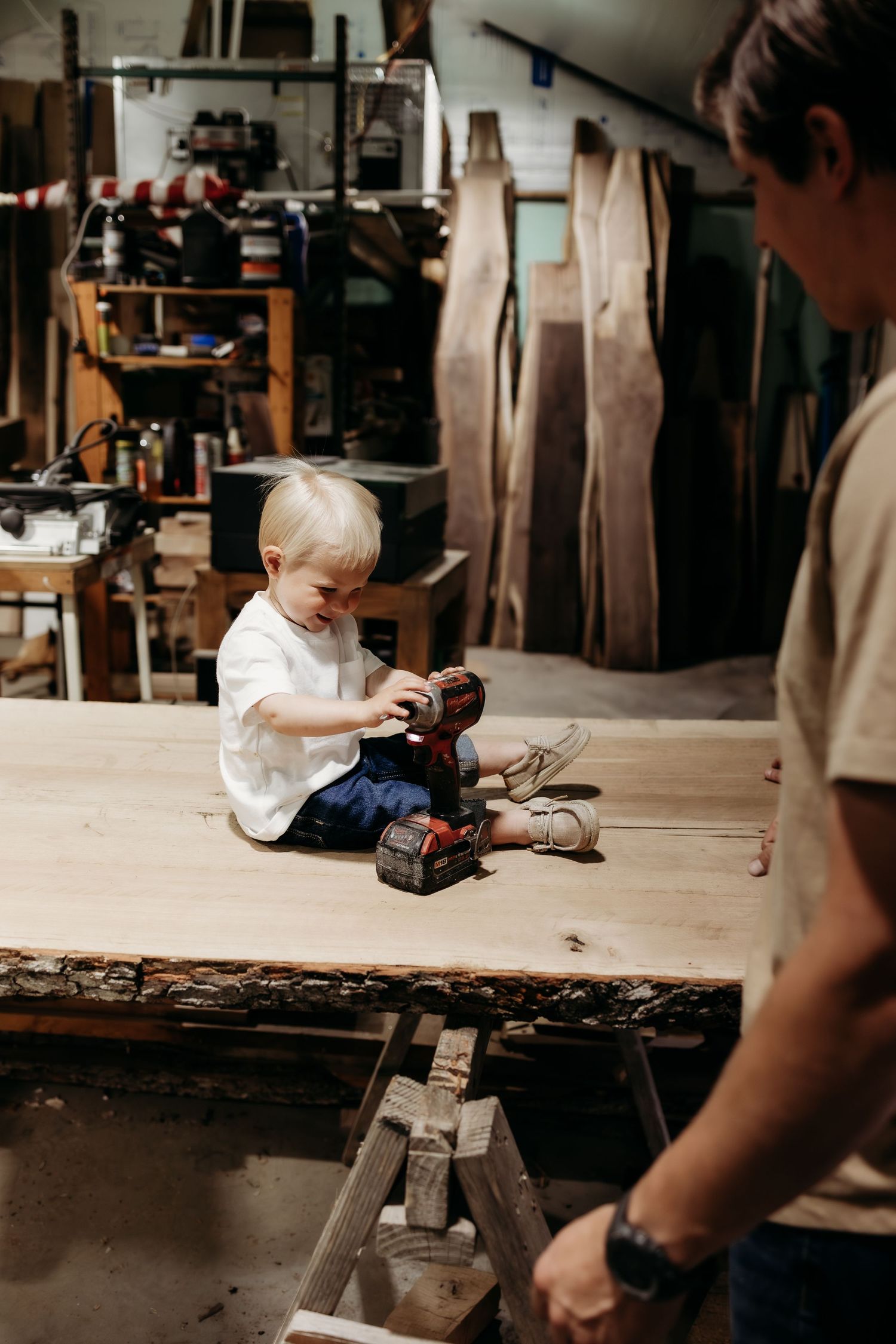 Toddler boy sitting on a workbench holding a power drill in a rustic woodworking shop.