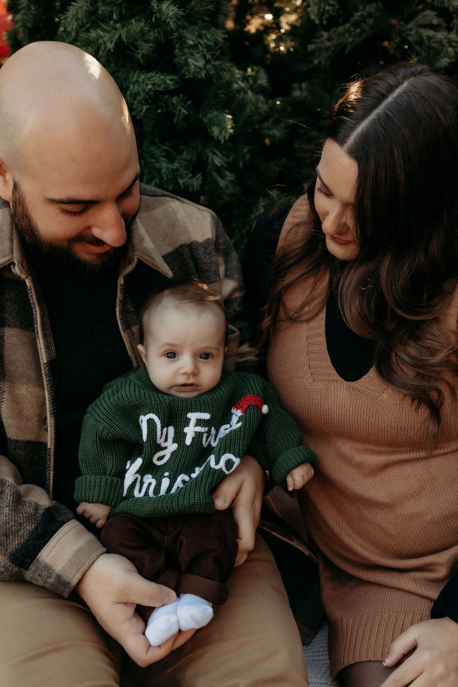 A young family poses in front of a Christmas tree, with their baby wearing a green My First Christmas sweater.