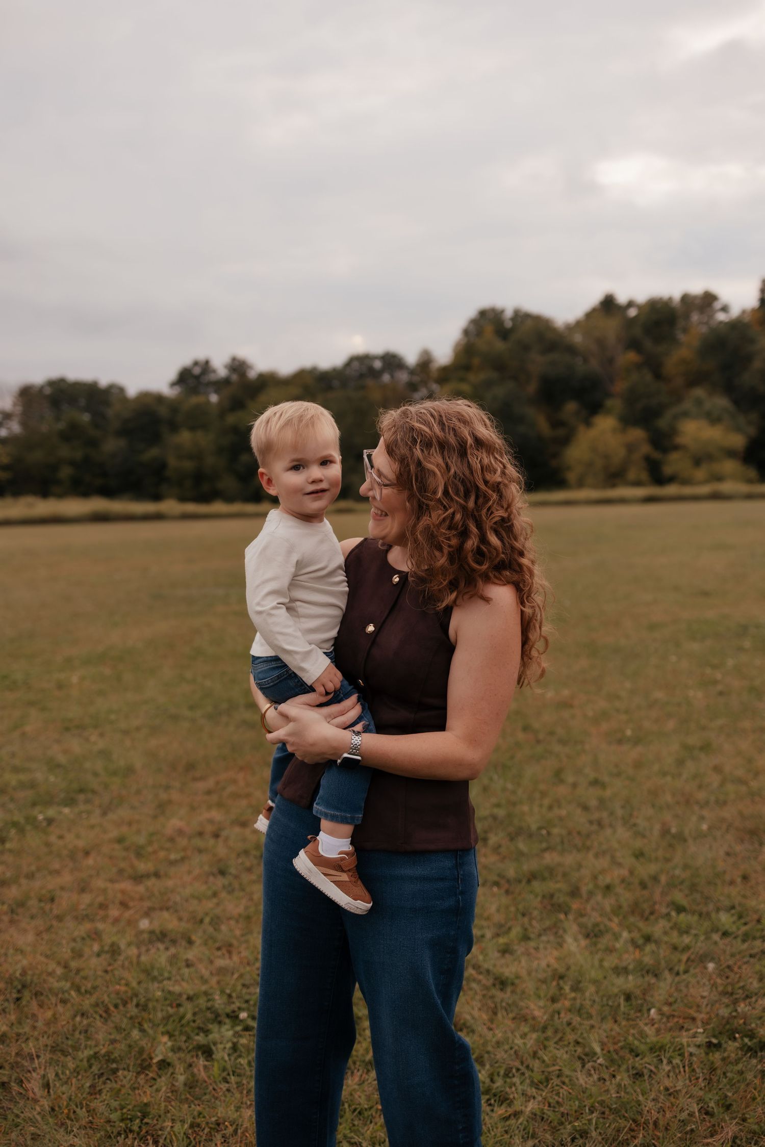 A curly-haired woman in a brown top holds a smiling toddler boy in a green field with trees in the background.