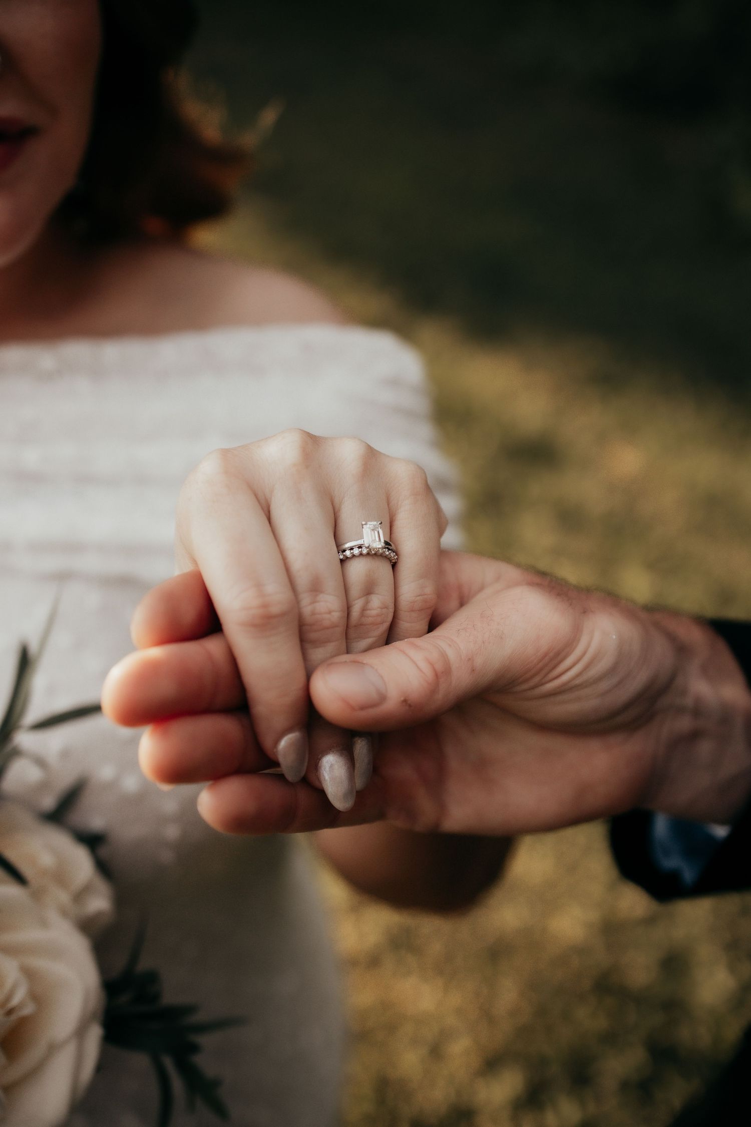 Bride wearing a diamond engagement ring holds hands with groom outdoors on their wedding day.