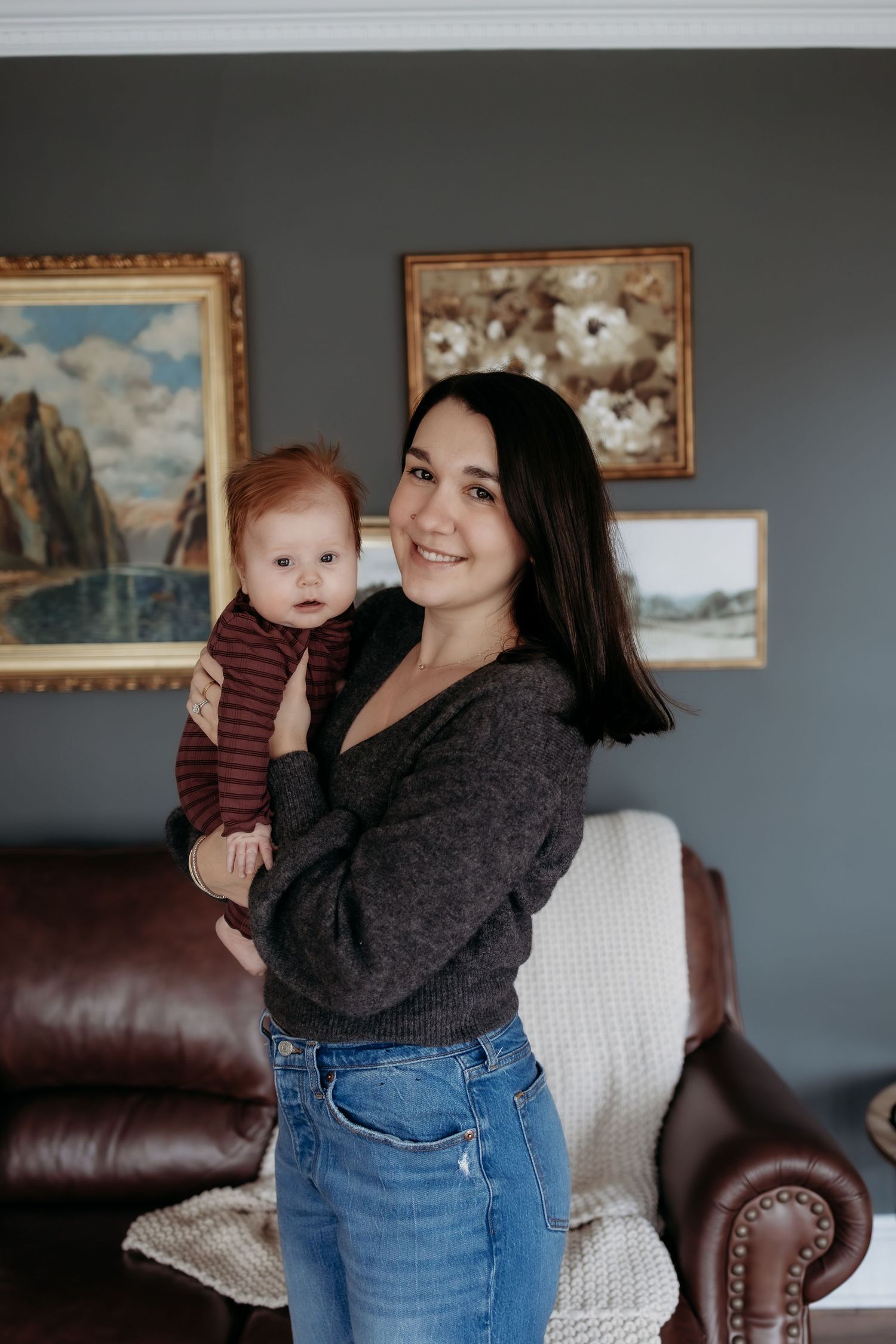 A smiling dark-haired woman holds a red-haired baby in a cozy living room with framed artwork on gray walls.