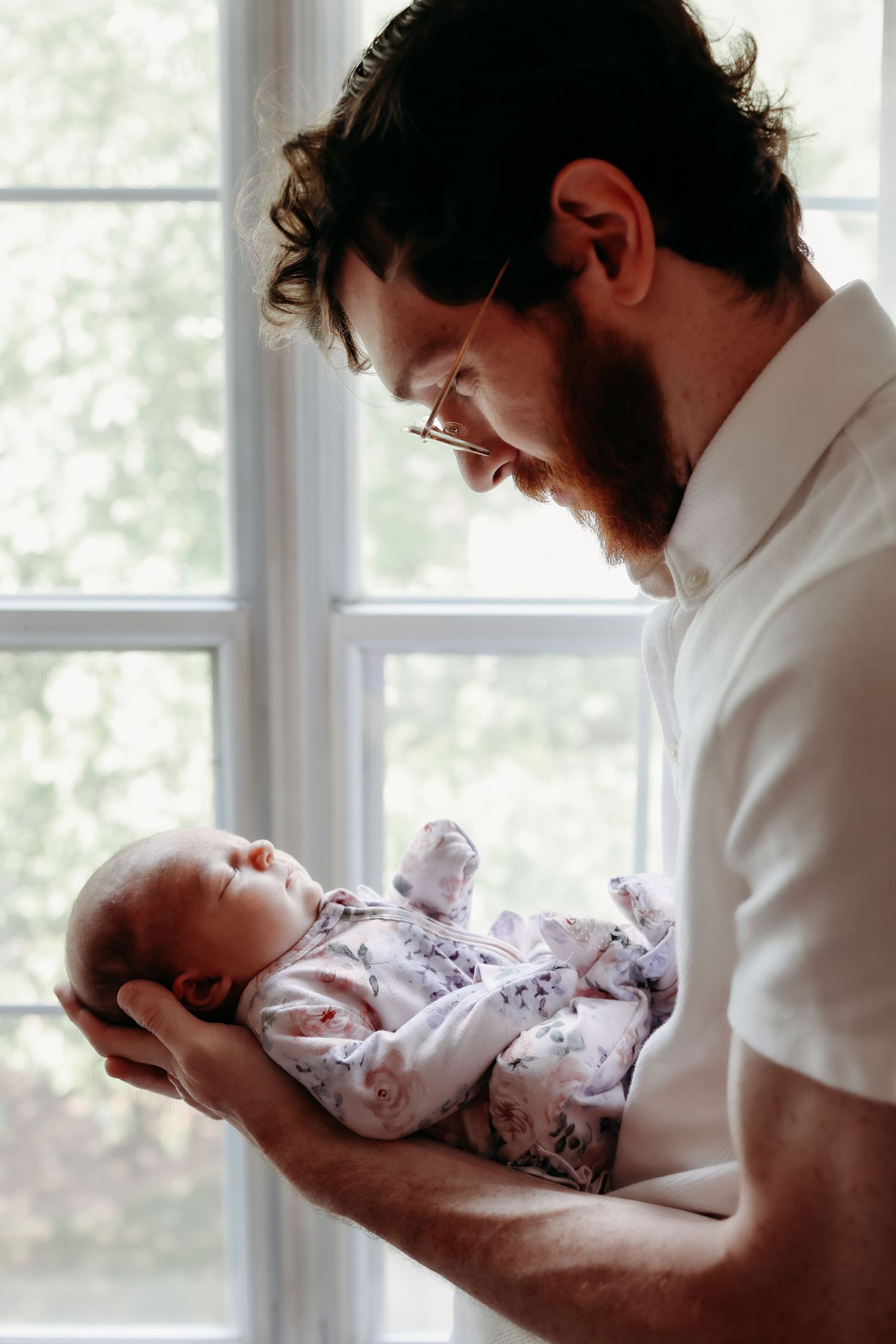 Bearded father with glasses lovingly gazing at sleeping newborn baby by bright window.