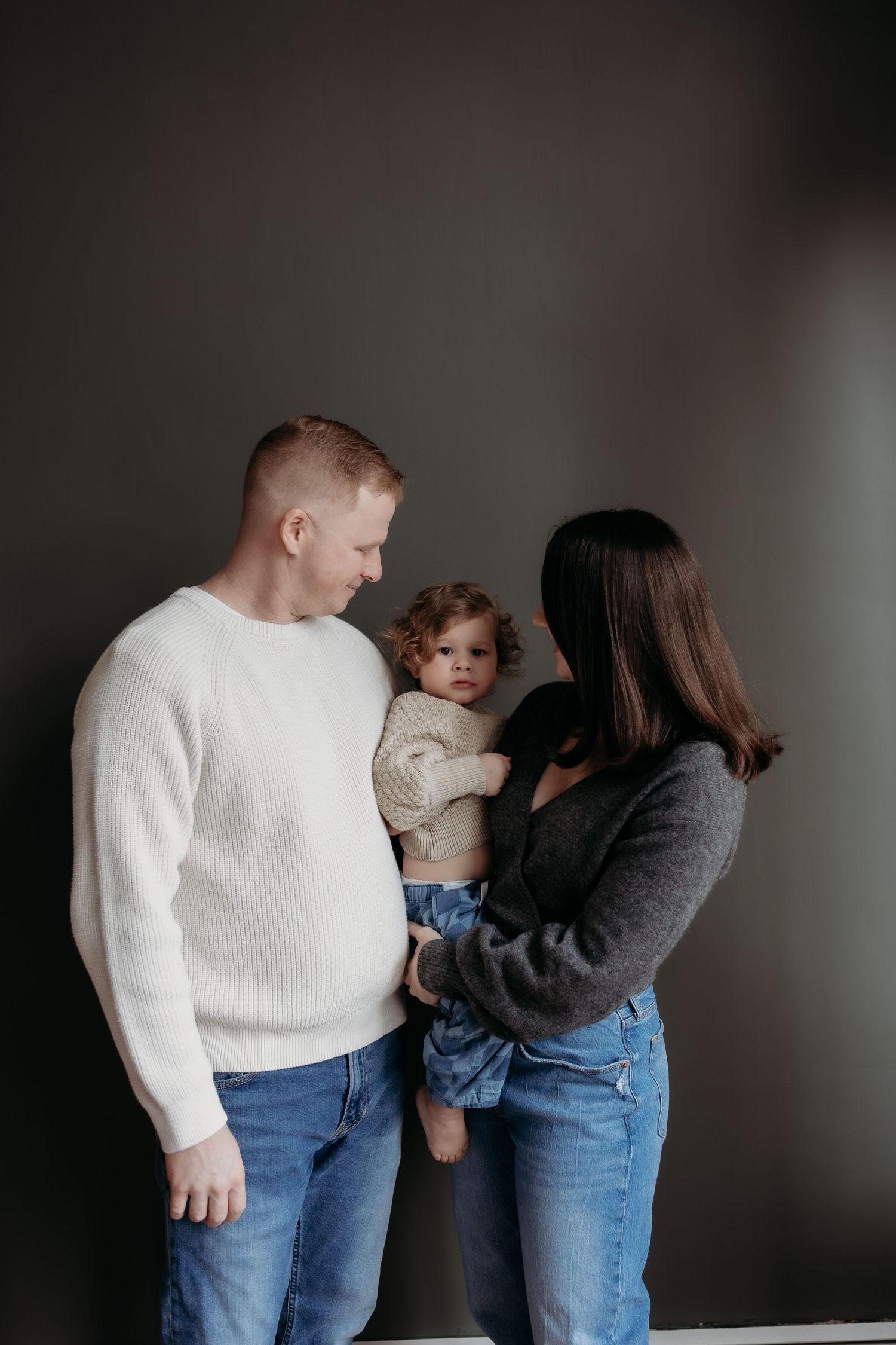 A young family of three poses indoors against a gray wall, parents gazing lovingly at their curly-haired toddler.