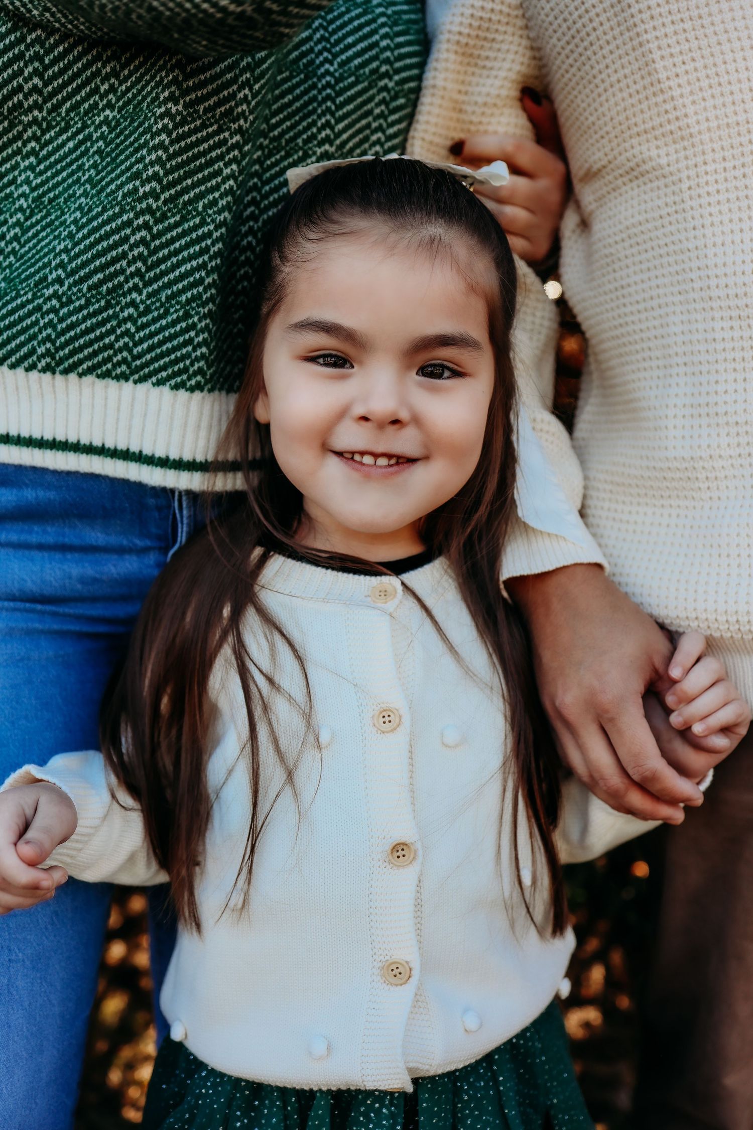 A smiling young girl with long dark hair in a white cardigan holds hands with adults in an outdoor autumn setting.