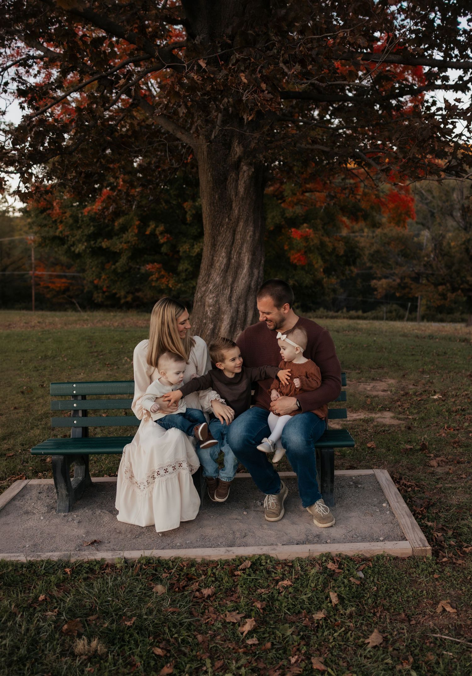 Family of four sitting on a green park bench under a large autumn tree with red and orange leaves, for a fall family portrait.