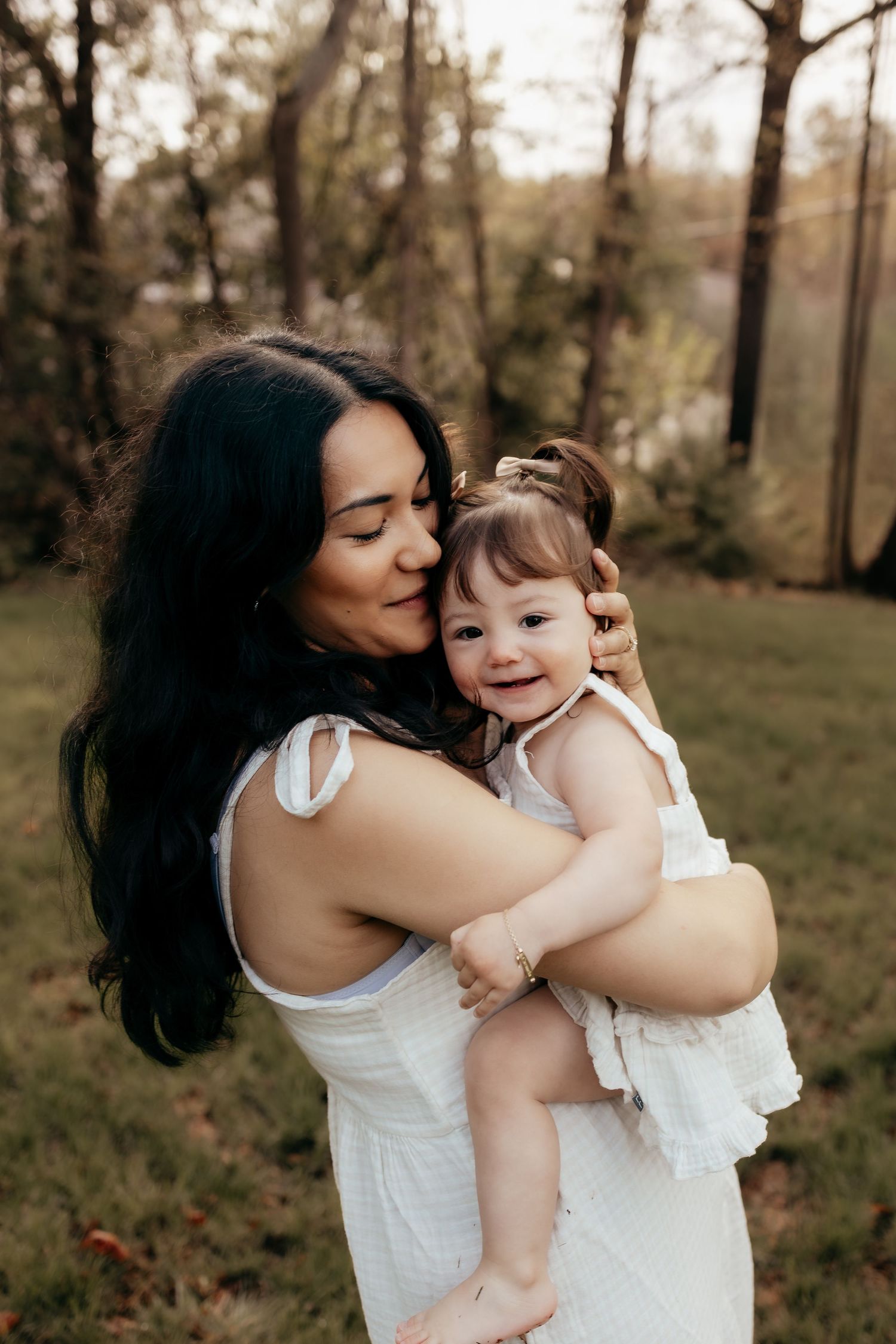 Dark-haired mother in white dress hugging smiling baby girl outdoors in a green park.