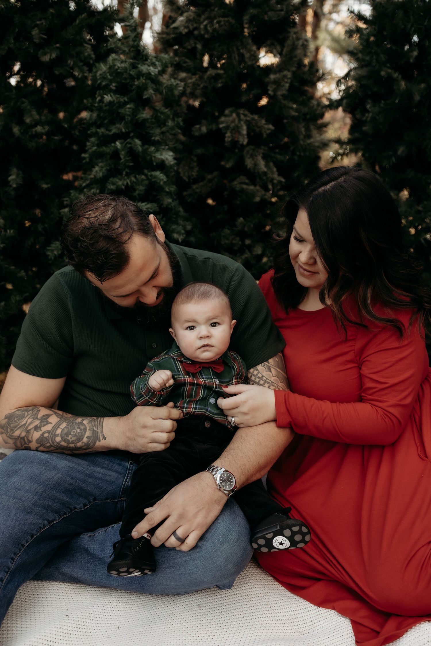 A young family sits outdoors near evergreen trees, parents lovingly gazing at their infant dressed in holiday plaid outfit.