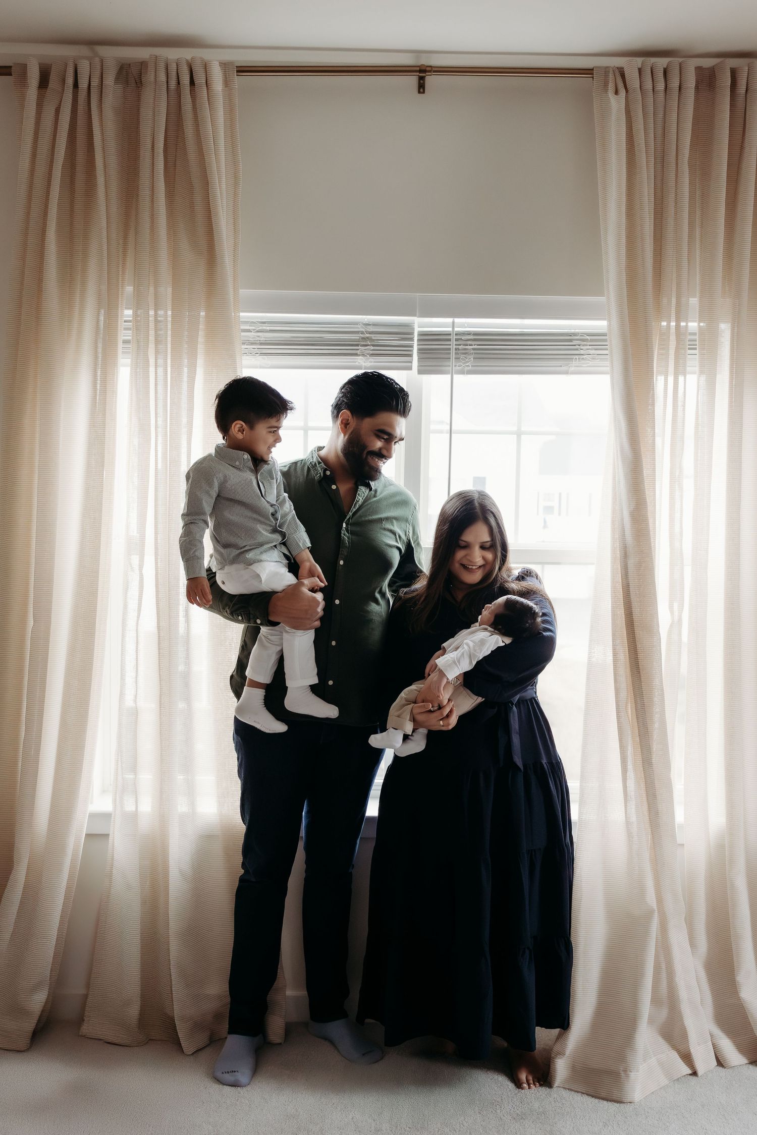 Family of four with toddler son and newborn baby posing before elegant linen curtains.