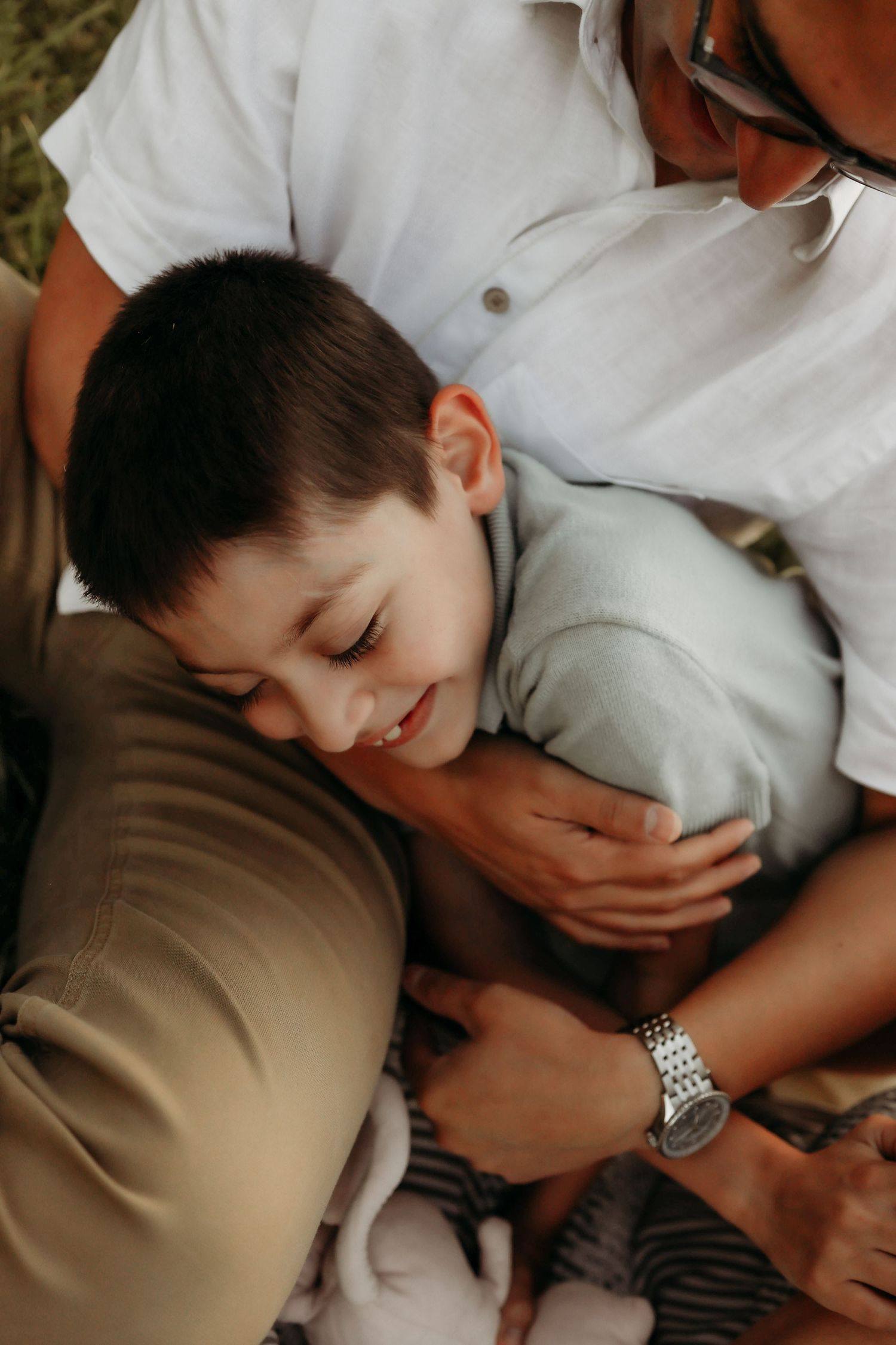 Young smiling boy cuddles between his parents in a warm, loving family embrace outdoors.