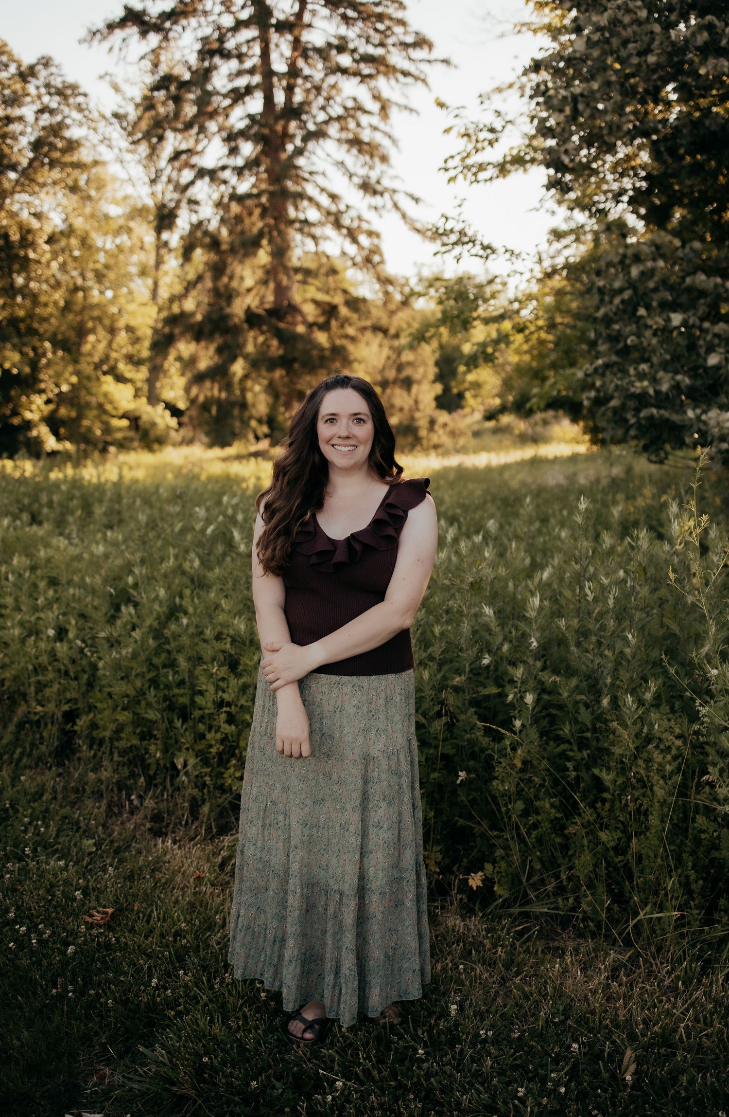 Smiling woman in dark ruffled top and maxi skirt stands in lush green field bathed in warm evening light.