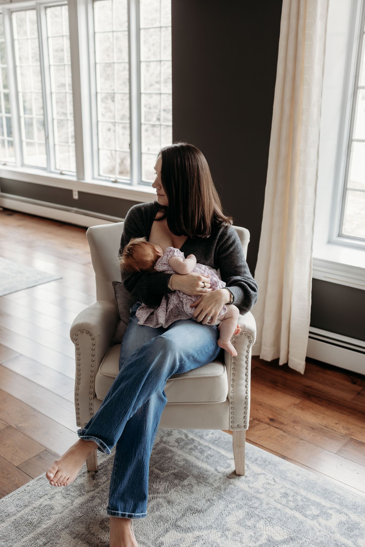Brunette mother sitting in white armchair breastfeeding infant near large sunlit windows.