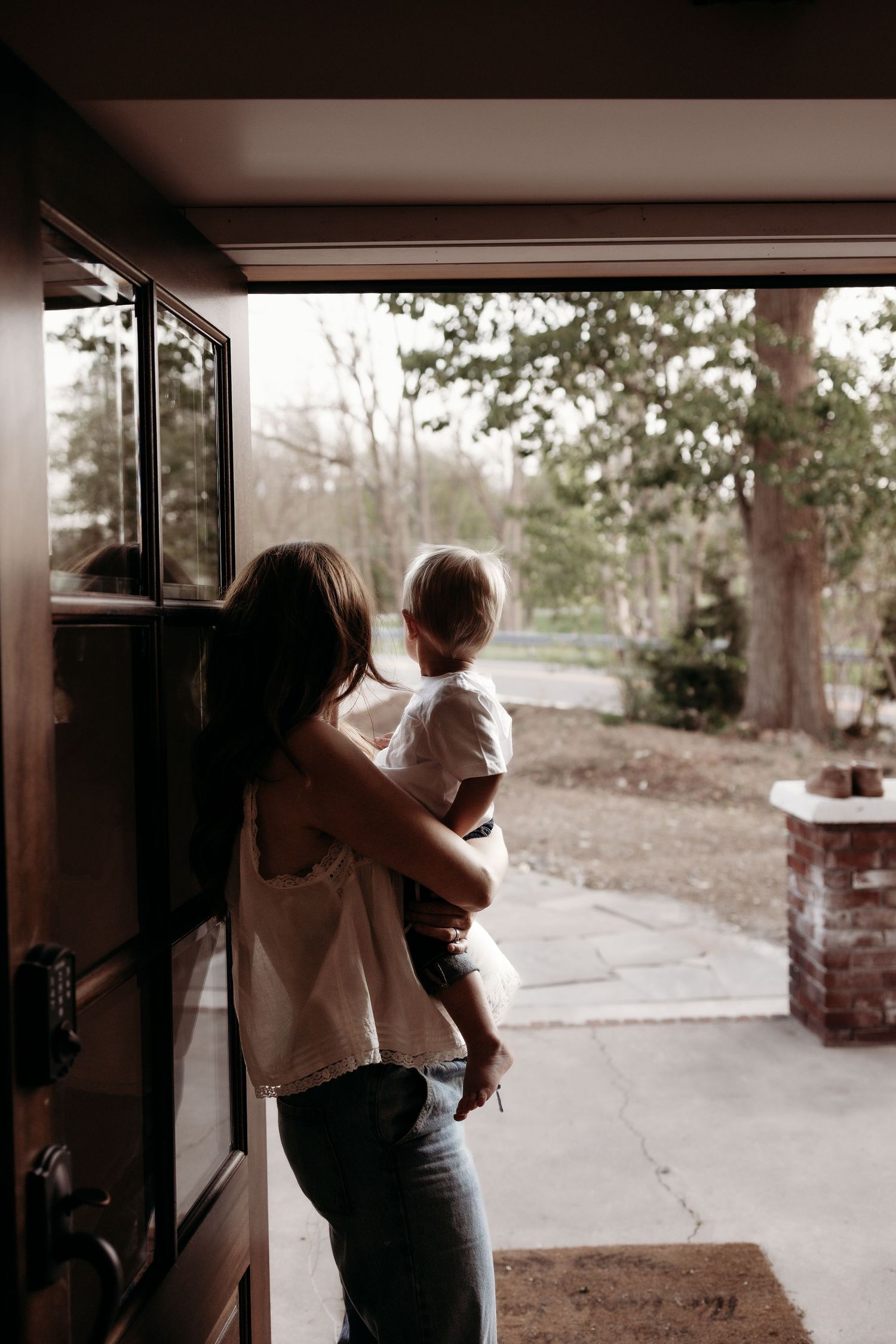 A mother holding her toddler while standing at an open front door, looking outside into the yard.