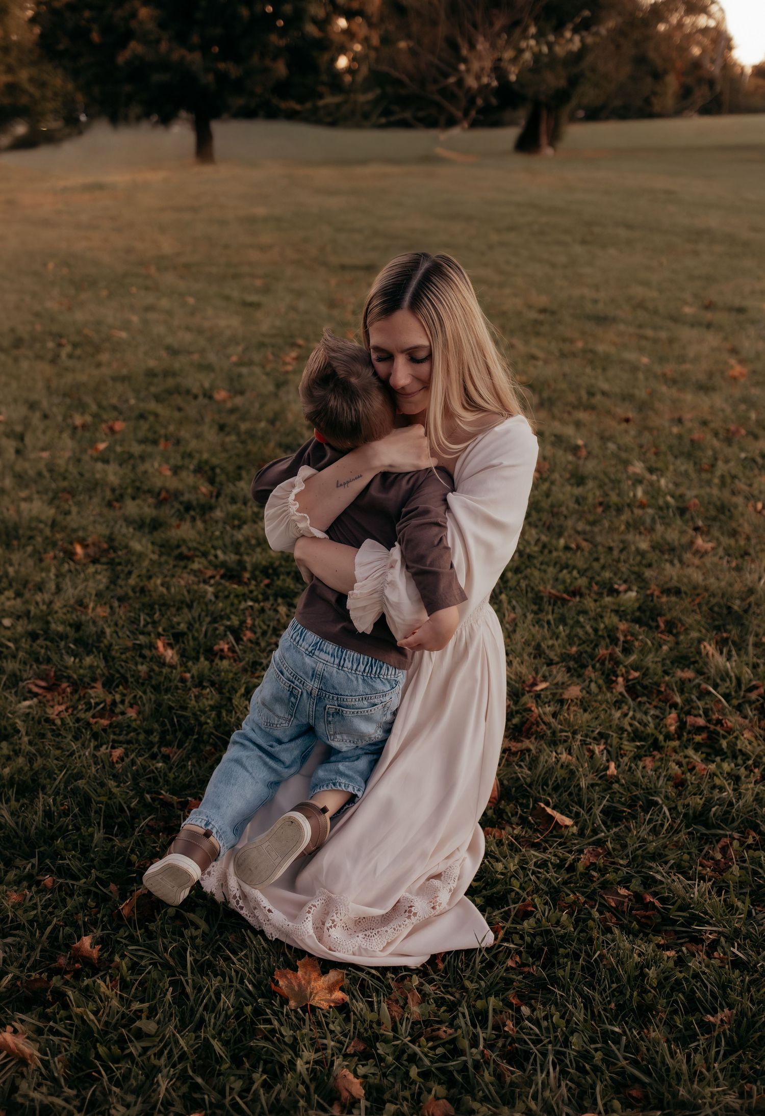 A woman in a cream dress kneels on autumn grass, lovingly hugging a toddler in jeans in a sunlit park with trees behind.