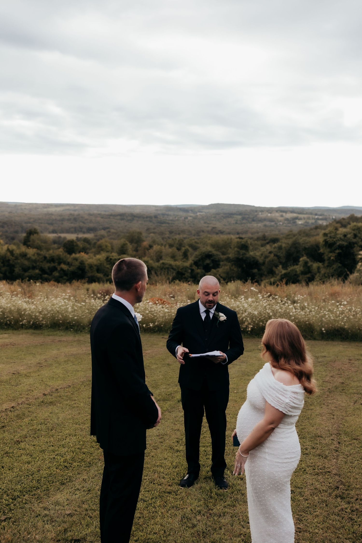 Outdoor wedding ceremony on a green hillside with bride, groom, and officiant reading vows under a cloudy sky.