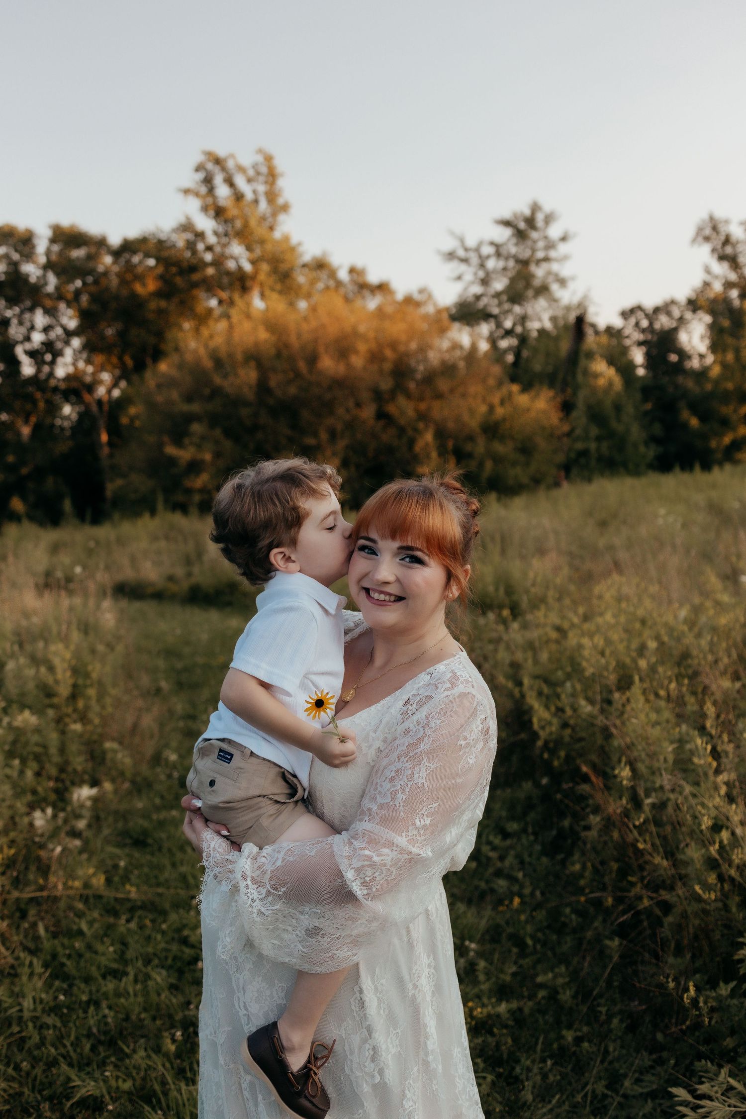 A red-haired woman in a white lace dress holds a young boy who kisses her cheek in a sunlit meadow.